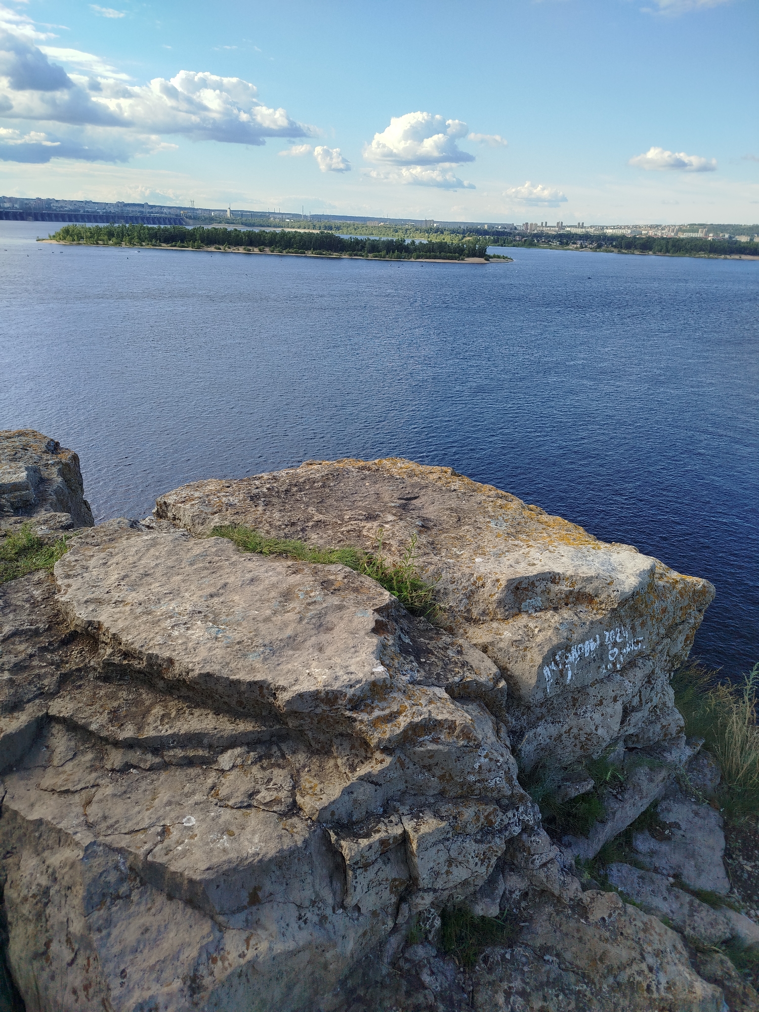 This is a landscape photograph taken from a high vantage point overlooking a wide, deep blue river. In the foreground, large, rough, grey rocks dominate the lower portion of the frame. These rocks are uneven, with sharp edges and some grassy patches growing between them. Some of the rocks have a yellowish lichen or moss growing on them, particularly on the right side. White graffiti is visible on the face of a rock on the right, which appears to include numbers and possibly some Cyrillic letters.
Beyond the rocky foreground, the expansive river stretches towards the horizon. On the far side of the river, a long, green, tree-lined island is visible, with a dam or structure running across the water behind it. Further in the distance, a city skyline with buildings and smokestacks can be seen along the riverbank, with more trees and greenery interspersed. The sky above is a bright blue, dotted with fluffy white clouds.
The lighting suggests it is daytime, likely late afternoon or early evening given the angle of the light casting shadows on the rocks. The weather appears to be clear and pleasant. The overall mood is one of natural beauty and serene observation. The location is likely Zhigulyovsk, Russia, given the context provided, which is known for its scenic views of the Volga River and the Zhiguli Mountains. There are no people clearly visible in the image, though some very small, dark shapes on the water might be boats. The graffiti on the rock is the only discernible text.