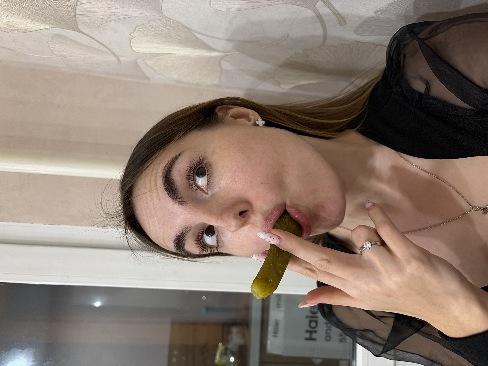 A woman is holding a pickle in her mouth while sitting indoors in what appears to be a residential setting. The background includes a wall with a floral-like pattern and a window with a blurry reflection of an object labeled "Haier". The woman is wearing a black semi-sheer top, a necklace, and a ring. Her nails are painted white with pink polka dots. There is no evidence of Izhevsk, Russia.