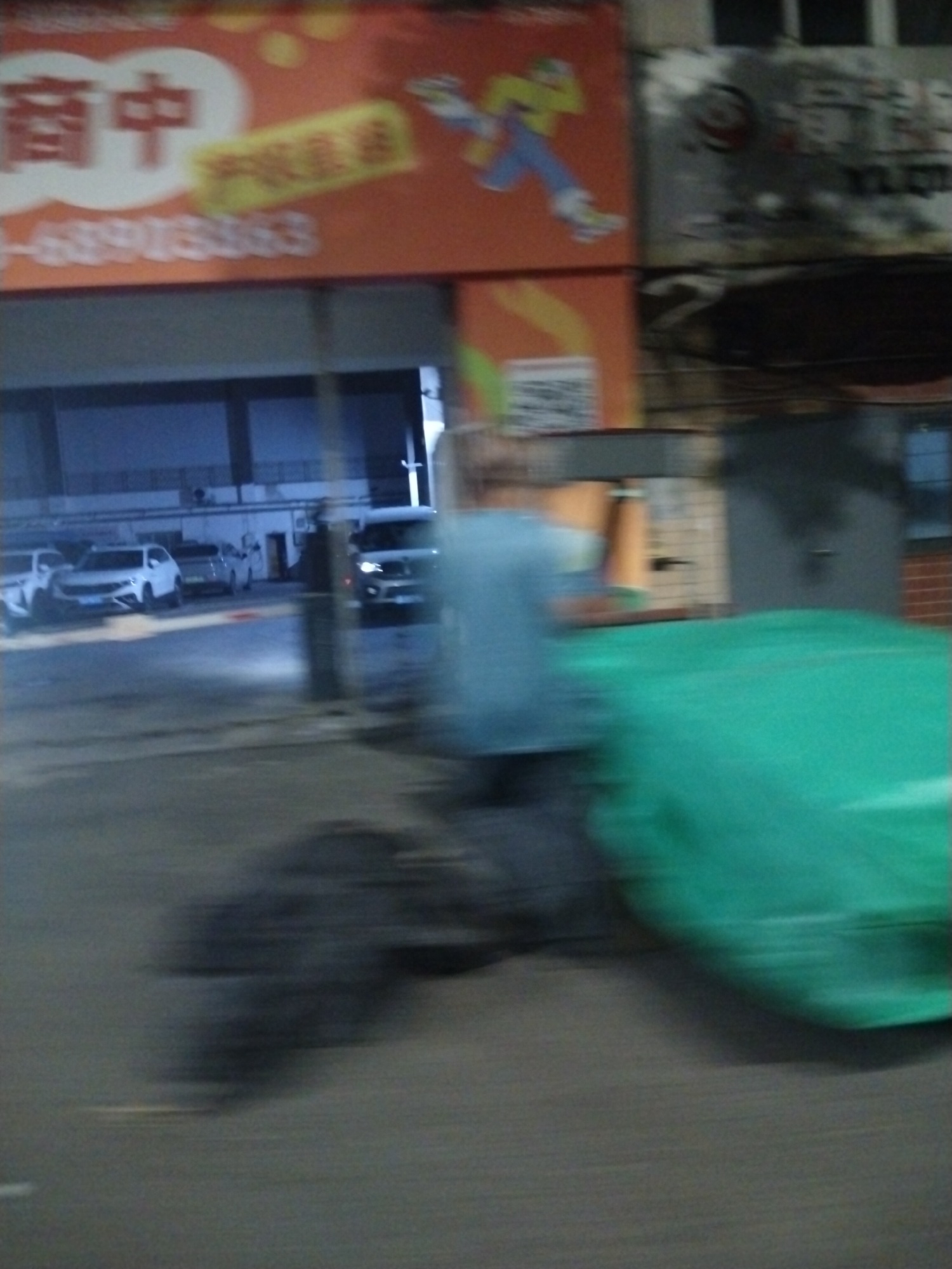 The image shows a street scene in Jiulongpo District, China. There is a building with an orange sign bearing Chinese characters and a cartoon figure. Several cars are parked inside a garage-like structure. A person is walking by with a green object. The photograph appears to be taken from a moving vehicle.