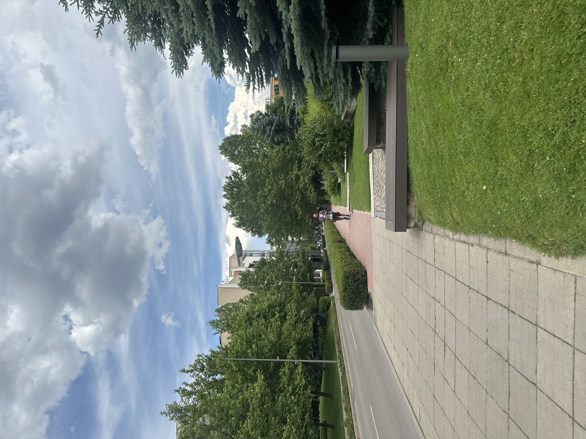 The image depicts a park or boulevard scene in Sofia, Bulgaria, on a cloudy day. A paved sidewalk runs alongside a road, bordered by a manicured green hedge and a grassy area. Tall evergreen trees and deciduous trees line the pathway, with one person walking on the sidewalk. In the background, modern buildings, including one with solar panels, rise above the trees. The sky is a mix of blue and white clouds, suggesting it is daytime. A bench with a dark wood finish and stone accents is visible near the trees. The overall impression is one of an urban green space. No visible text is present in the image.