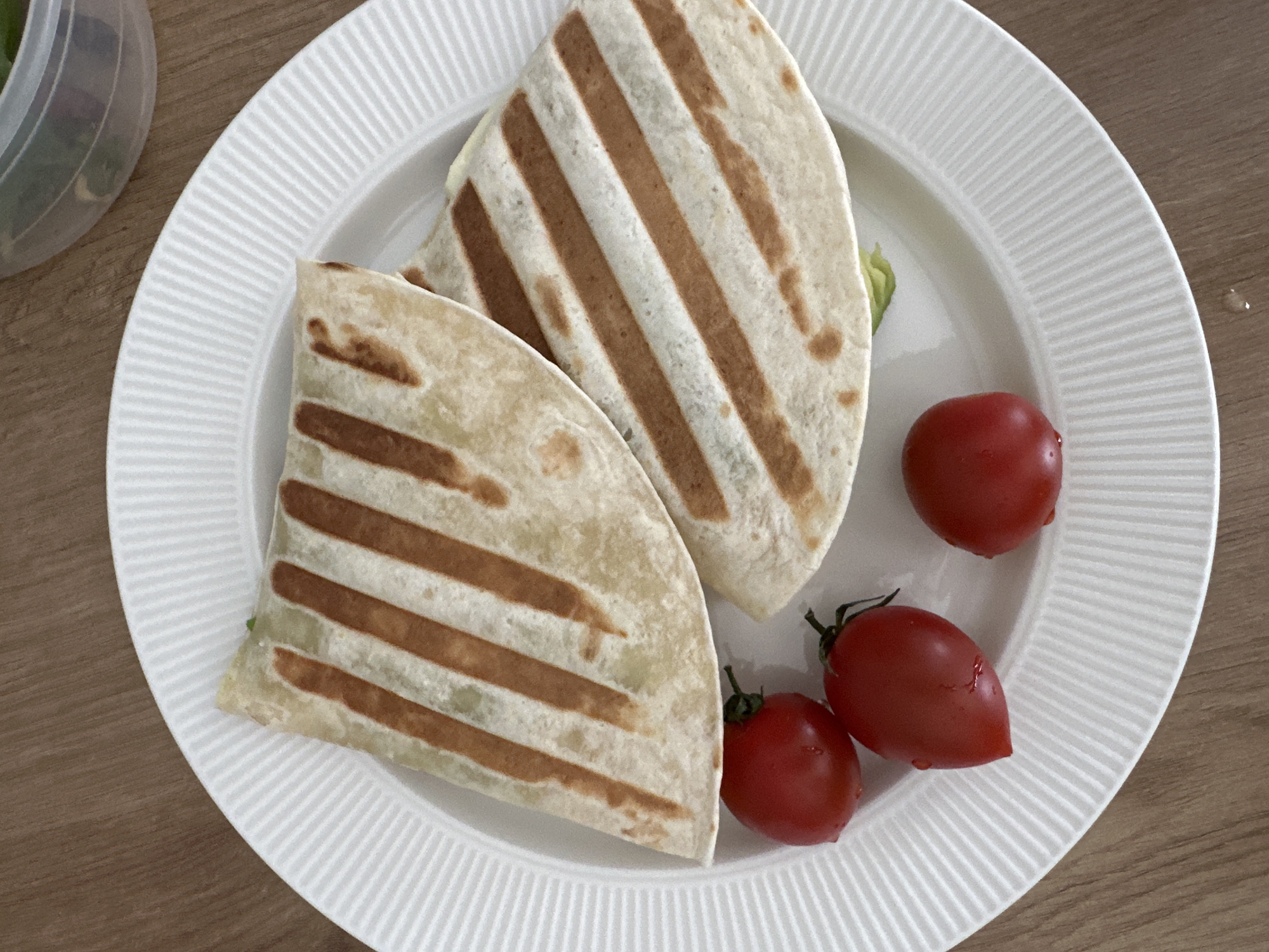 A top-down shot shows a white, ridged plate on a light brown wood-grain surface. The plate contains two halved wraps with distinct brown grill marks on the exterior. The wraps are filled with greens and other ingredients. Three red cherry tomatoes are arranged on the plate alongside the wraps. A partially visible container with green contents is located at the top left of the frame. The location is not discernible from the image.