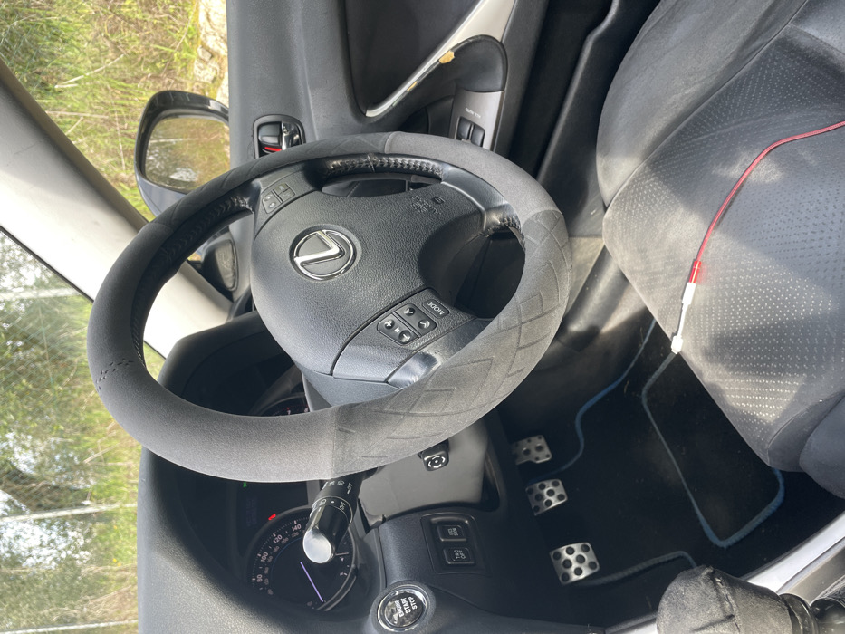 The scene depicts the interior of a Lexus vehicle, specifically the driver's area, consistent with an outdoor setting in Monsummano Terme, Italy. A black steering wheel with a silver Lexus emblem and integrated audio/mode controls is partially covered by a two-tone gray fabric cover featuring a diamond stitch pattern. The instrument cluster shows indicator lights illuminated and the speedometer needle at zero. To the left of the steering column, an 'ENGINE START STOP' button is visible. Below, three metal-trimmed pedals (clutch, brake, accelerator) rest on a dark floor mat with blue piping. A manual gear shifter with a black fabric boot is present in the lower left. The perforated black driver's seat and a portion of the front passenger seat are visible. A red and white audio cable lies on the front passenger seat. The right-side interior door panel features silver trim and a red door lock indicator. Through the vehicle's windows, a sunlit exterior displays green foliage and a chain-link fence. No specific landmarks or precise street location within Monsummano Terme are discernible from the visual evidence.
