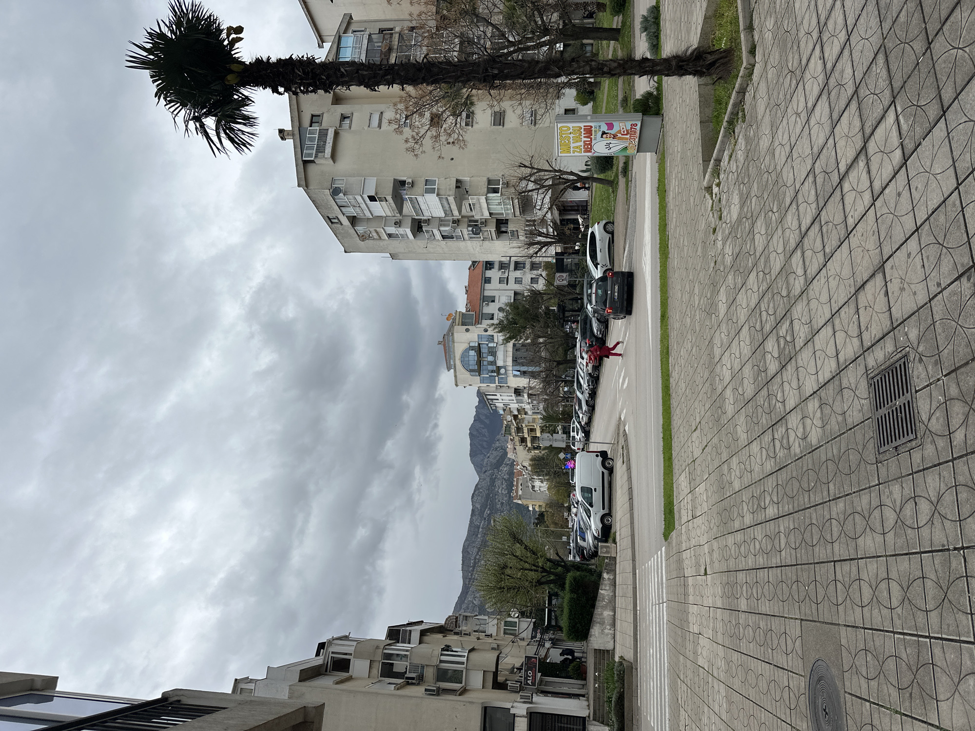 The image captures a street scene in Bar, Montenegro, showcasing a blend of urban and natural elements. The perspective is from the sidewalk, which is paved with geometric-patterned tiles. A palm tree stands prominently on the left, reaching towards an overcast sky. Buildings, primarily apartment blocks, line the street, varying in height and architectural style, with some featuring balconies and air conditioning units.
A row of parked cars occupies the right side of the street, leading towards a backdrop of distant mountains shrouded in clouds. A pedestrian crossing is visible in the foreground. A billboard featuring a person in wedding attire is visible near one of the buildings. A person wearing a red jacket is lying on the street between two cars. The overall atmosphere suggests a cloudy day in a residential area of Bar.