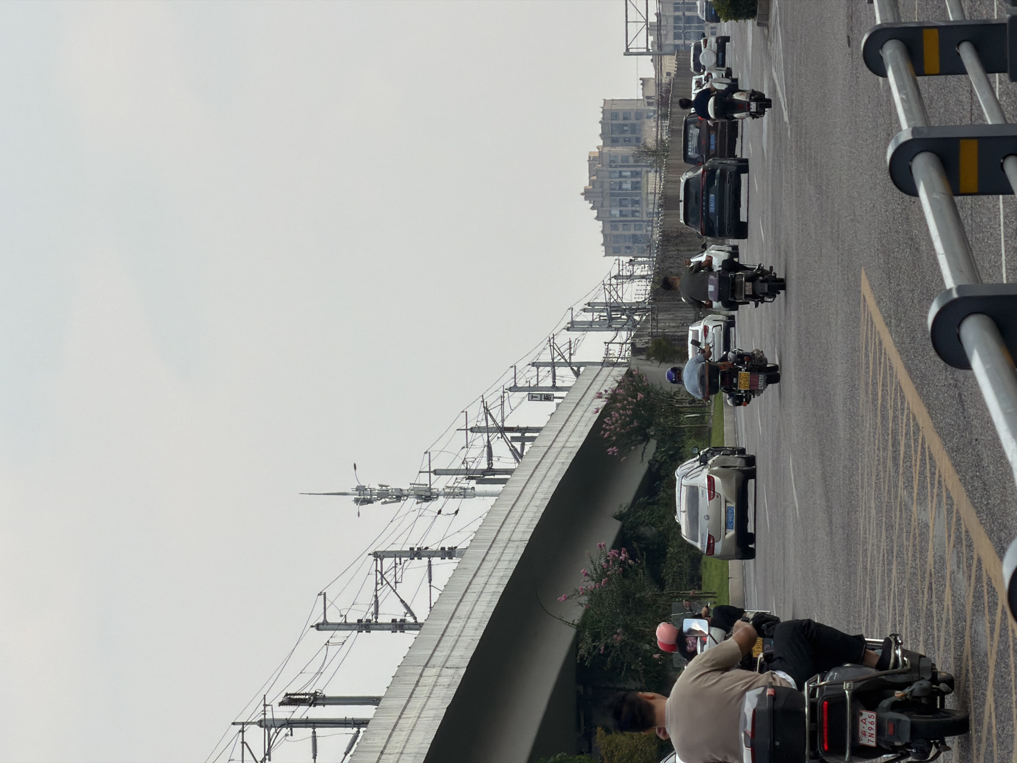 The image depicts a street scene in Jiangjin District, Chongqing, China, under an overcast sky. In the foreground, a person on a dark-colored electric scooter, wearing a beige shirt and black pants, is seen from behind. The scooter has a black storage box and a license plate displaying "A 7N965". To the left, another scooter is partially visible.
Further down the street, several vehicles are in motion or stopped. A silver sedan with a license plate reading "C G9592" is traveling in the same direction. Behind it, a black SUV and a brown SUV are also on the road. Several other motorbikes and scooters are present, some with riders.
Overhead, a large elevated concrete structure, likely a bridge or overpass, spans across the scene. It is supported by numerous pylons and is equipped with extensive electrical wiring and transmission towers. In the background, several multi-story buildings are visible, including a large, tan-colored building with a distinctive stepped facade. Green foliage and bushes line the sides of the road and the area beneath the elevated structure. The road surface is asphalt with yellow lane markings.