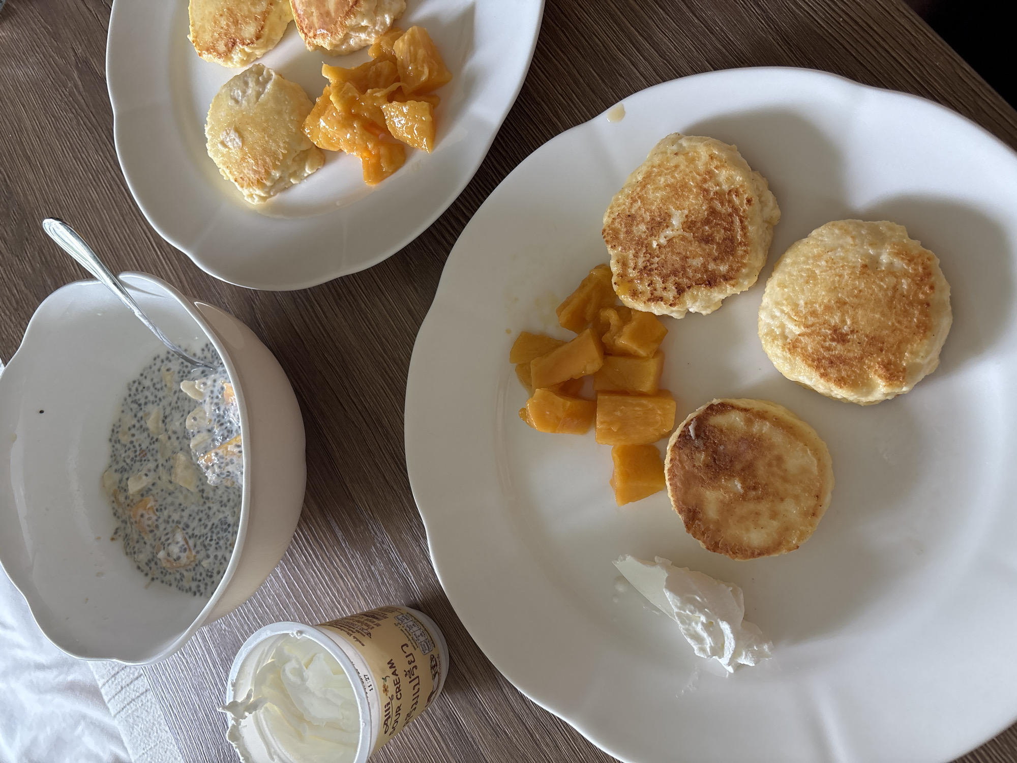 This overhead shot captures a breakfast or brunch spread laid out on a warm-toned wooden table, likely in Choeng Thale, Thailand. The scene features two white plates with scalloped edges, each holding several small, round, golden-brown pancakes or fritters, possibly *syrniki* due to their appearance and accompanying condiments. Alongside these, portions of diced yellow-orange fruit, resembling cooked mango or papaya, are present on both plates.
The larger plate on the right displays four pancakes, fruit, and a dollop of white cream, likely sour cream, at its bottom right. To the lower left of the frame, a white bowl, resting on a white cloth or napkin, contains a creamy greyish mixture speckled with black chia seeds and small fruit pieces, indicative of chia pudding or overnight oats. A silver spoon rests inside the bowl. Adjacent to the bowl, a plastic tub, partially visible, contains more of the white cream. Visible text on the tub clearly reads "SOUR CREAM" in English, accompanied by Thai script (likely "นมเปรี้ยว" meaning sour milk/cream) and a numerical detail "11.27", possibly a date or time.
The arrangement suggests a peaceful, prepared meal for one or two people, possibly in the morning or early afternoon. No individuals are visible, allowing the focus to remain on the neatly presented food items. The combination of Western-style pancakes/fritters with tropical fruit and sour cream, along with the chia pudding, points to a health-conscious and culturally blended meal, fitting the specified location context.