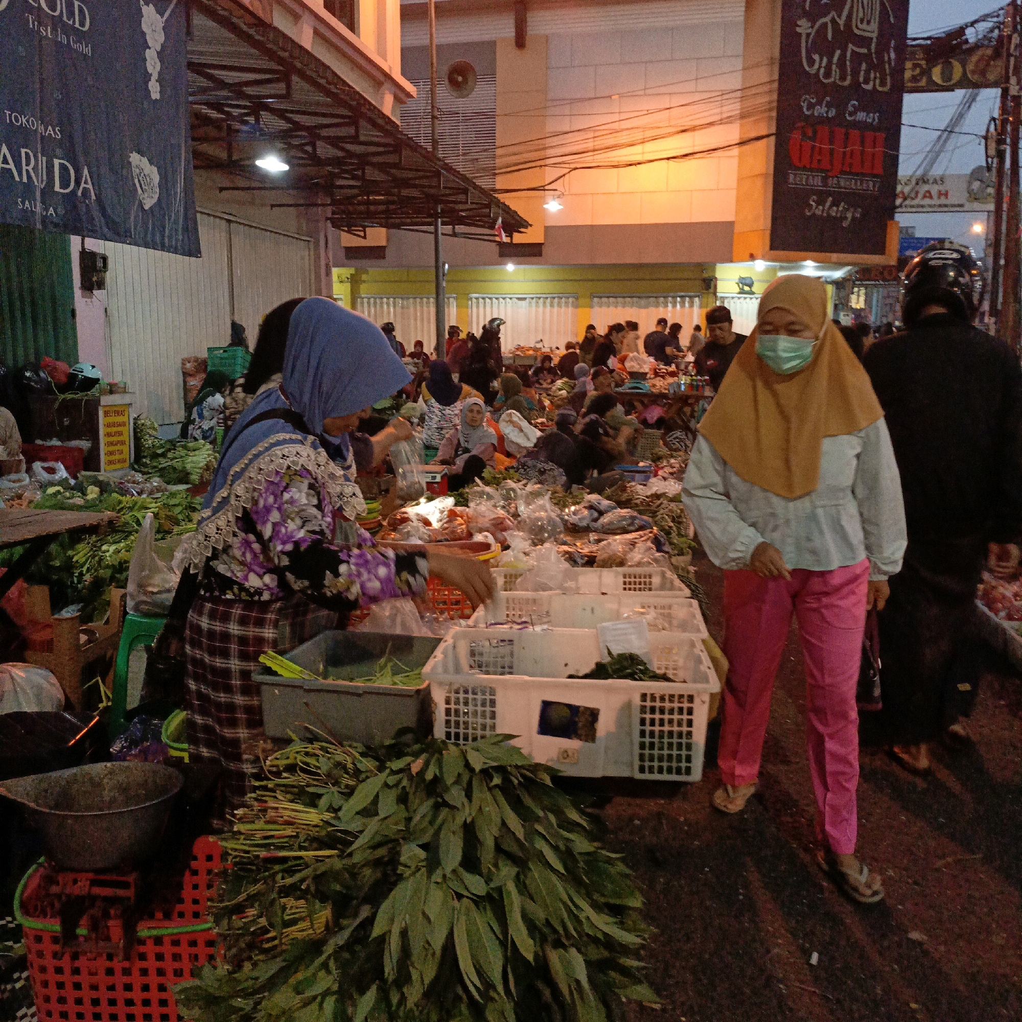 The image depicts a bustling street market scene, likely in Salatiga, Indonesia, characterized by an array of vendors and shoppers. In the foreground, a woman wearing a blue hijab and a floral patterned top, along with a plaid skirt, is actively engaged with produce, appearing to sort or package items. To her right, another woman in a tan hijab and a light blue shirt is seen wearing a face mask and pink pants, holding a dark bag. The market is filled with stalls displaying various goods, prominently featuring fresh produce such as leafy greens arranged in piles and in baskets. Several white plastic crates are visible, some containing more produce. In the background, numerous people are milling about, some interacting with vendors, while others are walking. Structures with awnings and signs indicate commercial establishments. The lighting suggests it is either dusk or dawn, with artificial lights illuminating parts of the scene. The overall atmosphere is one of lively commerce and community activity.