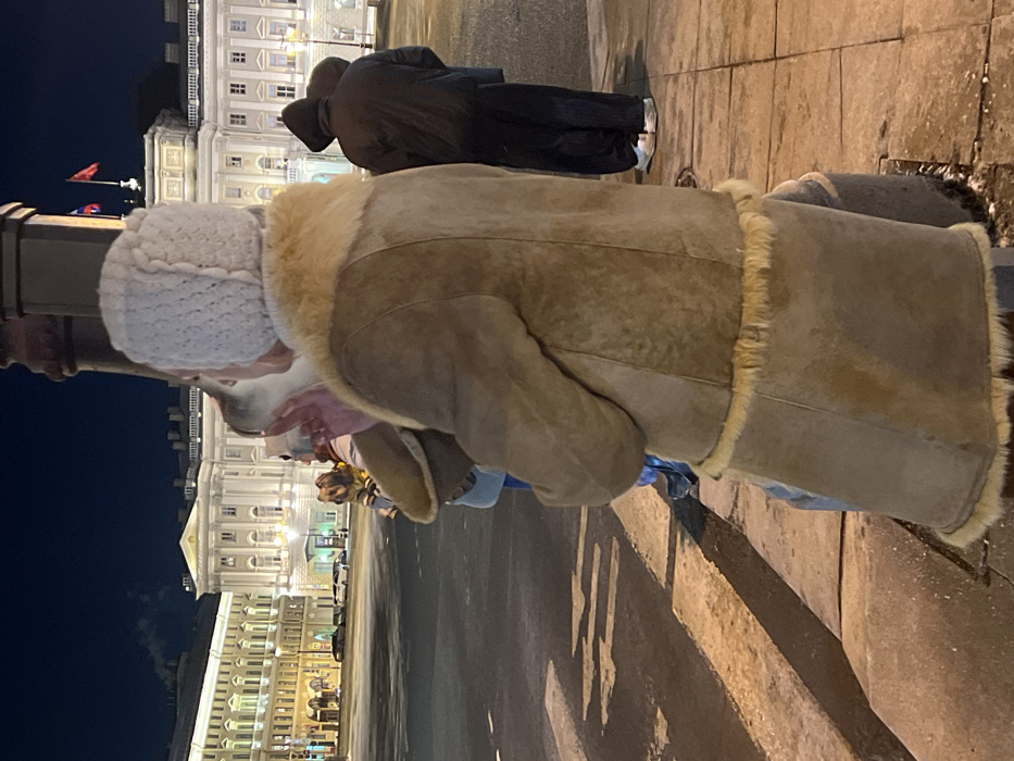 The image, rotated 90 degrees clockwise, captures a nighttime scene on a city street or plaza. The dominant figure in the foreground is a person viewed from behind, slightly turned to their left. They are wearing a light-colored, possibly off-white, knitted hat with a textured pattern. Their torso is covered by a warm, light brown or tan shearling-lined coat or jacket, featuring visible cream-colored fur trim on the collar and along the edges. The person appears to be bending slightly or looking down, interacting with a small dog whose head is visible just below their chin. The dog has light-colored fur on its head. Further down, near the person's feet, a small, dark-furred dog, possibly brown and black, is visible, suggesting it's the full body of the dog the person is interacting with, likely on a leash.
Further back in the midground, another person stands, also viewed from behind. They are wearing a dark, likely black, hooded winter coat and white sneakers.
The background features grand, multi-story buildings with classical architectural elements, such as columns and ornate window frames. These buildings are brightly illuminated, creating a warm glow against the dark night sky. To the left of the main buildings, two red flags are displayed on flagpoles. The ground is paved, appearing somewhat reflective in areas, possibly from moisture, and strong shadows are cast by unseen light sources, indicating streetlights or building lights. The overall atmosphere suggests a cold evening in a historic urban environment. No text is visible in the image.