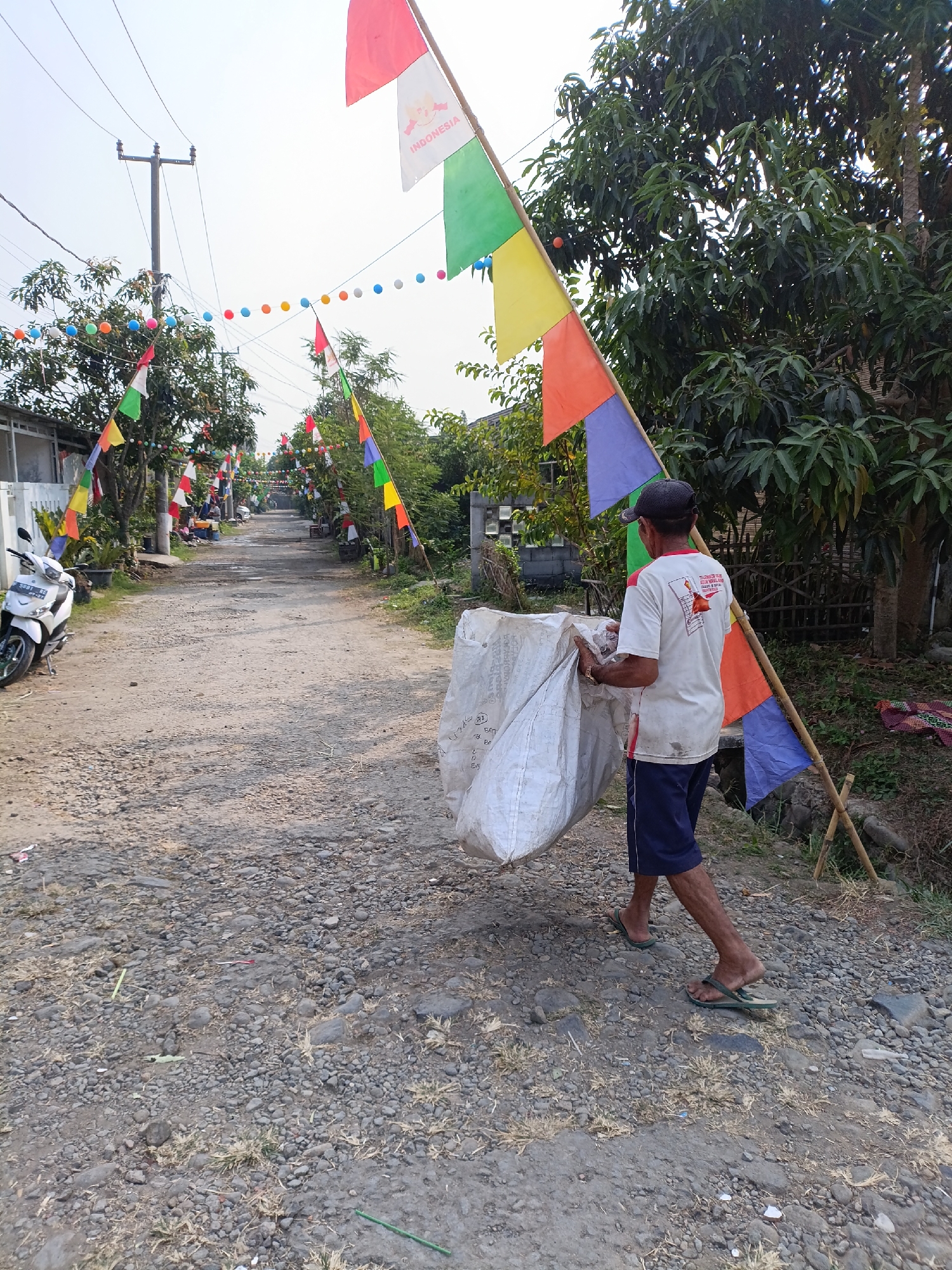 A man walks down a gravel road in Cariu, Indonesia, carrying a large white sack over his shoulder. He is wearing a white t-shirt with a graphic on the back, dark blue shorts, and flip-flops. The road is decorated with colorful flags and balloons strung across overhead wires. A white motorcycle is parked on the left side of the road, and trees and houses are visible in the background. The scene appears to be set during daylight hours.