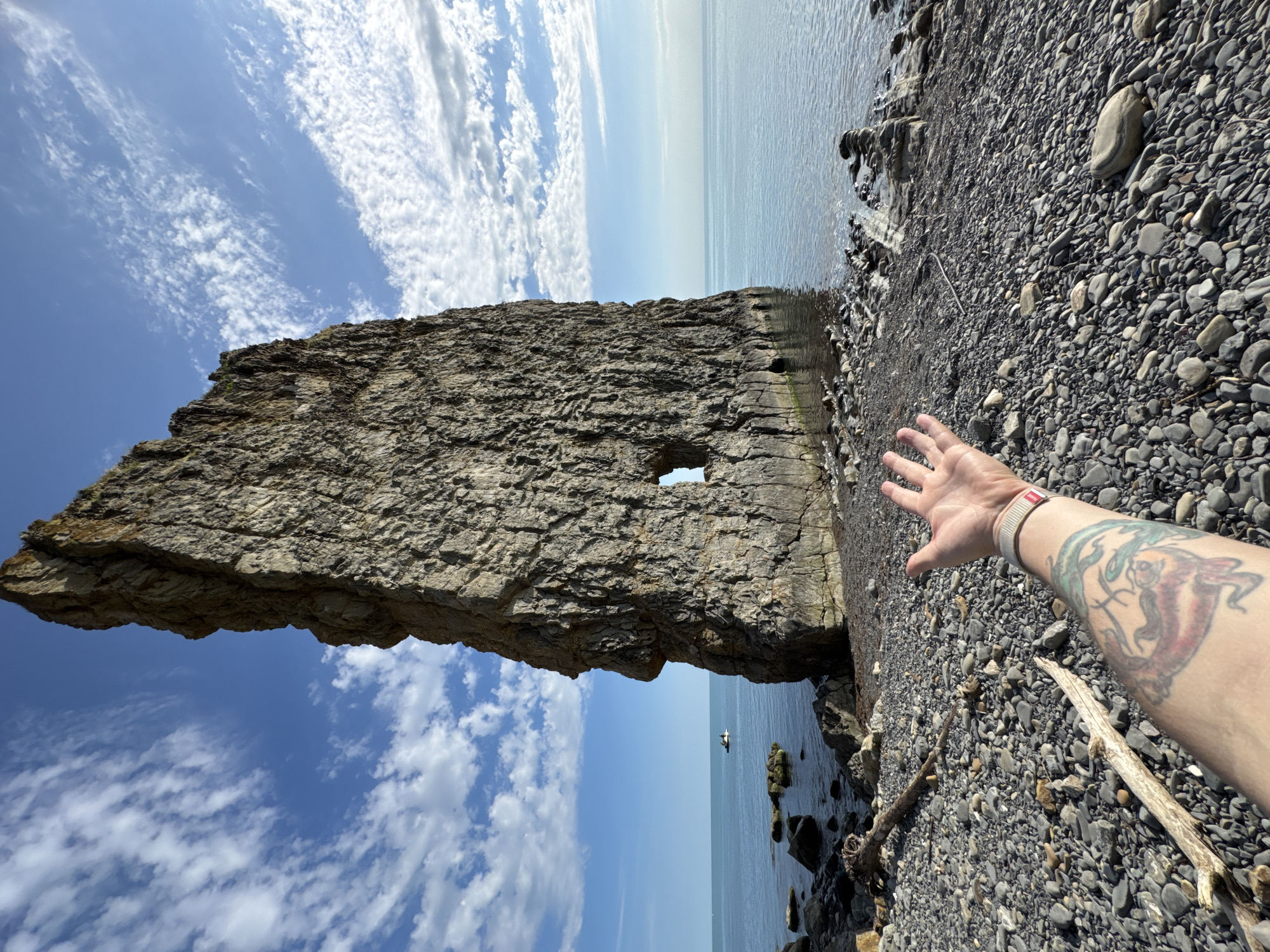 This media file presents a vibrant coastal landscape featuring a distinctive natural rock formation under a clear, sunny sky, likely mid-morning or afternoon.
The central subject is a colossal, tall, and remarkably flat rock formation, resembling a sheer wall or pillar, dominating the left side of the frame. This impressive geological marvel, recognized as Sail Rock (Parus Rock) in Divnomorskoe, Russia, rises dramatically from the water's edge and features a prominent, perfectly rectangular opening or archway carved near its base. Through this natural arch and stretching into the distance is the tranquil, blue-green sea, with a small boat or paddleboard and its occupant visible far out on the water.
Above the rock and the sea, the sky is a bright blue, adorned with scattered wispy white cirrus clouds and some larger cumulus formations, hinting at fair weather. The foreground on the right consists of a pebble beach, densely covered with smooth, grey and black stones of various sizes, with a few pieces of driftwood scattered among them.
In the lower right corner, a person's tanned right arm and open hand extend into the frame, reaching towards the magnificent rock formation or the sea. The arm displays a vibrant, colorful tattoo, possibly depicting a stylized marine creature, and a light-colored wristband. This outstretched hand suggests a personal interaction with the stunning natural environment, perhaps as part of a photograph or to convey a sense of scale. No visible text is present in the image.