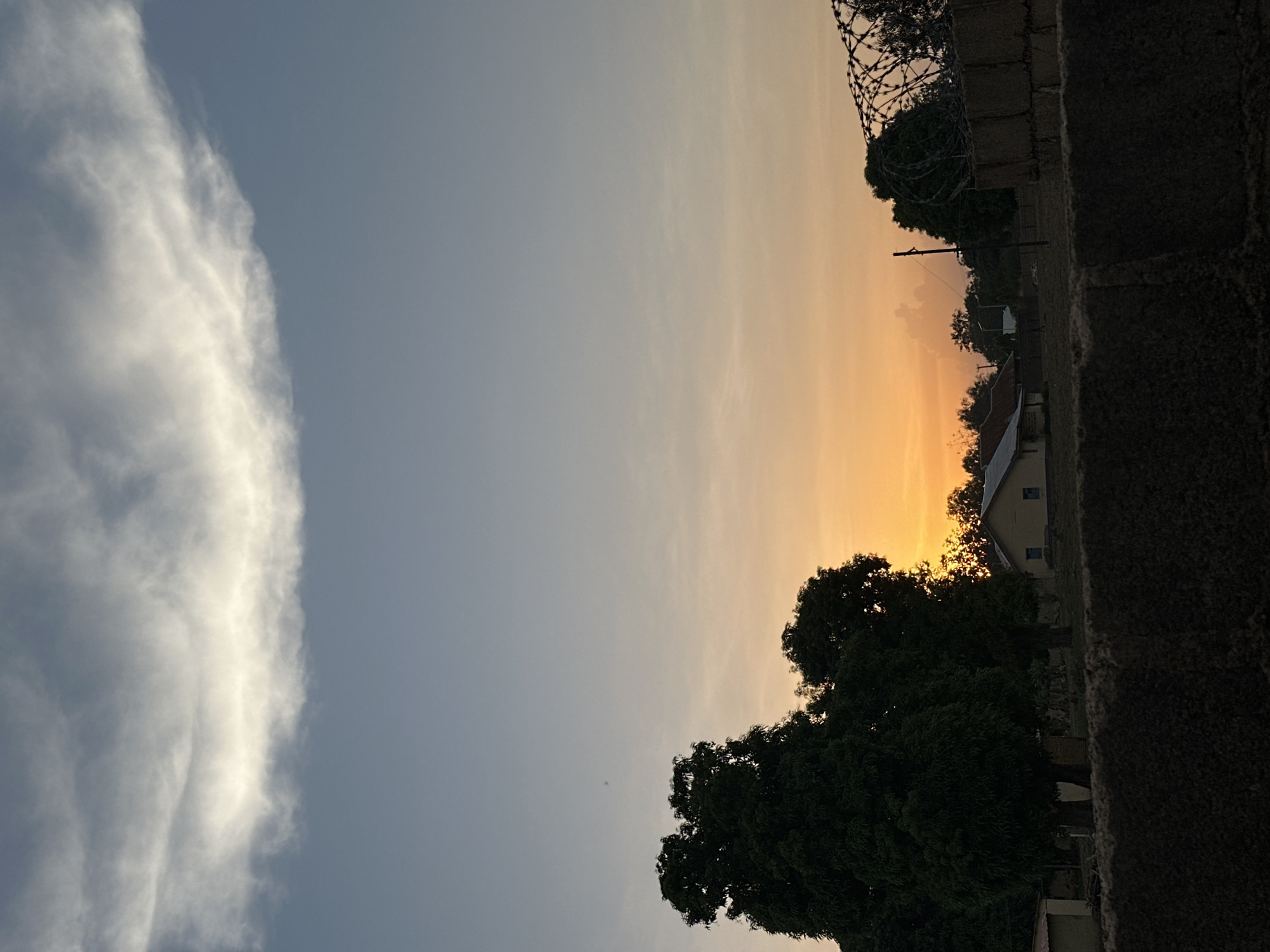 The image depicts an outdoor scene in Bukuru, Nigeria, at what appears to be sunset or sunrise, given the orange glow in the sky. In the sky on the left side of the image is a long, thin cloud formation. The sky transitions from blue to orange. The right side of the image includes a dark brick or concrete wall topped with what appears to be barbed wire. The silhouette of trees are visible against the orange sky, with a building partially visible behind them. The building has a sloping roof and visible windows.