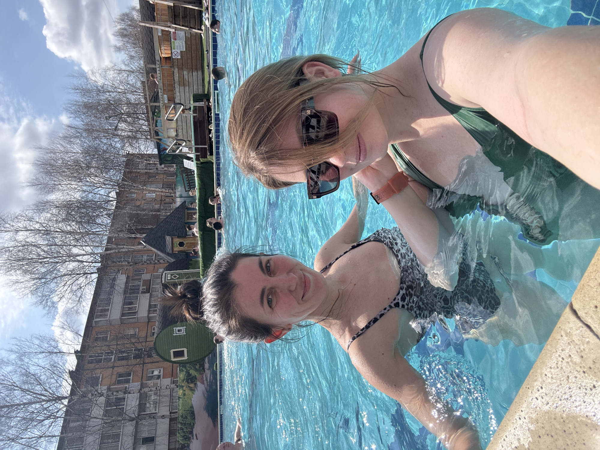 Two women are in a swimming pool. One woman is wearing sunglasses and a green bathing suit, and the other is wearing a black and white leopard print bathing suit. The pool is located in Votkinsk, Russia, as evidenced by the urban landscape in the background. Other people are also in the pool.