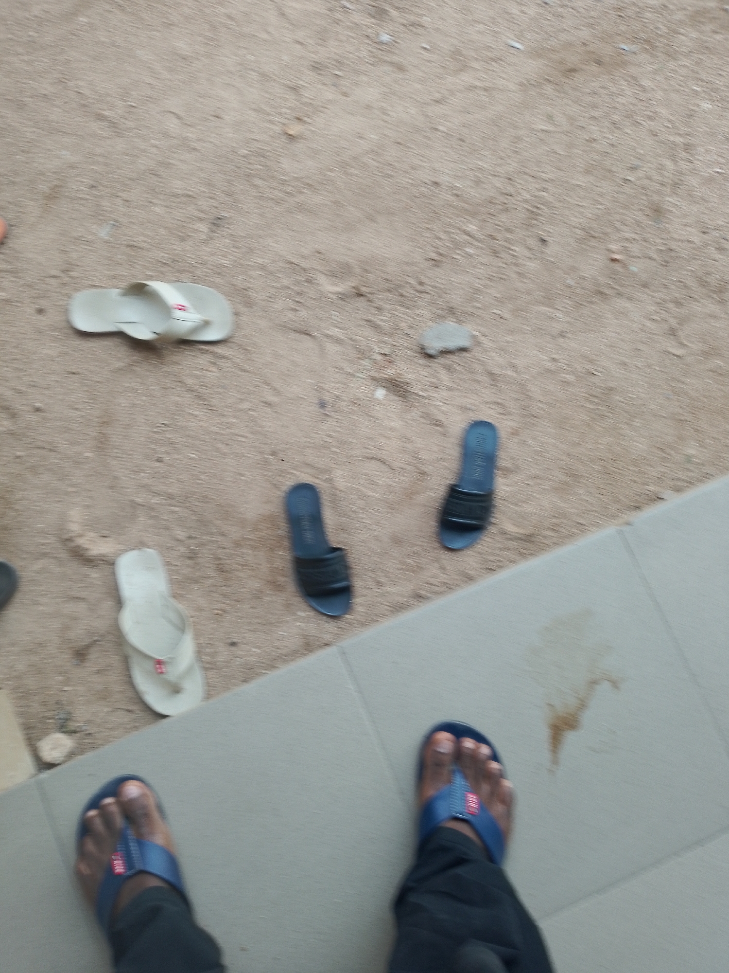 The image depicts a ground-level view of several pairs of flip-flops scattered on sandy and tiled surfaces. In the upper left, a light-colored flip-flop is visible on the sand. Further to the right on the sand, another light-colored flip-flop lies near a small grey stone. Below these, a pair of dark blue flip-flops are positioned. In the foreground, two feet wearing blue flip-flops are visible, standing on a tiled surface. One foot is more prominent, showing dark skin and toes. The tiled area also has a light brown stain. The overall environment appears to be outdoors, possibly a pathway or courtyard. The location is identified as Unknown, Unknown.
