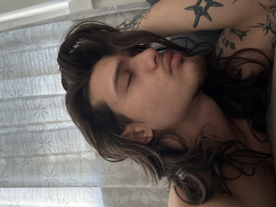 The image displays a young adult individual, appearing male, with long, dark, wavy hair, lying in a relaxed position with eyes closed and lips slightly puckered. A light goatee is present on the chin, and several moles or freckles are visible on the right cheek. A small, silver-colored hoop earring is in the left earlobe. The individual's left arm is bent, with the forearm or hand partially supporting the head. Multiple dark-ink tattoos are visible on the left arm and shoulder, including a prominent barbed wire or crown of thorns design on the upper arm, and various abstract patterns. The individual appears shirtless. The background features white, translucent curtains patterned with repeating floral or starburst designs, indicating an indoor setting with a window. The immediate surface beneath the individual's head and arm is dark, appearing to be soft bedding or a pillow. The scene is observed indoors, likely within a bedroom environment, in Sofia, Bulgaria.