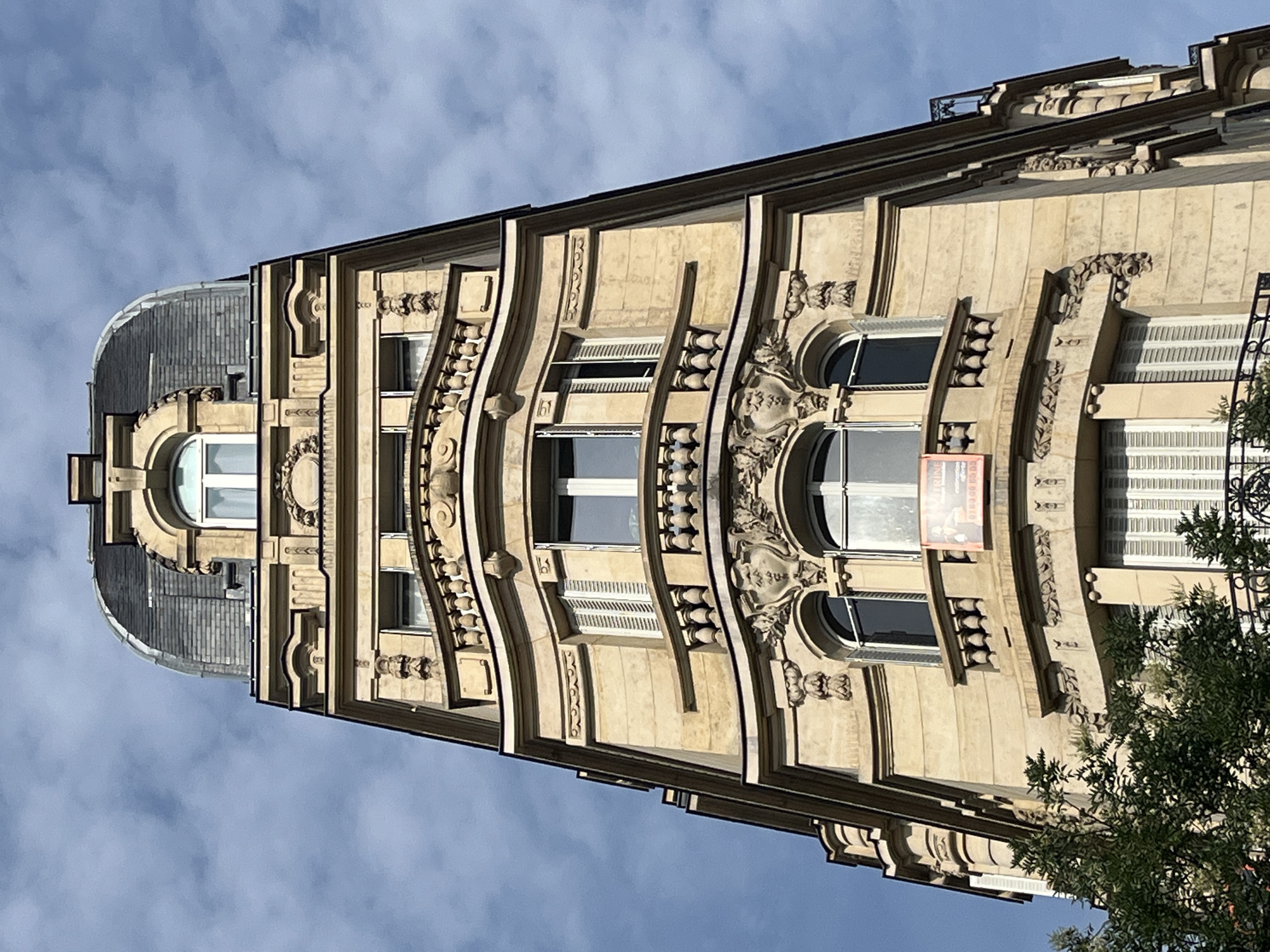 The image is a low-angle, full shot of the upper floors of a tall, ornate building in Paris, France. The building has a distinctive Mansard roof covered in dark grey slate tiles and topped with a finial. The facade is constructed of light-colored stone and features decorative elements such as carved moldings, balustrades, and arched windows. Several windows are visible, some with closed white shutters. A small orange sign is attached to a balustrade on one of the floors. The sky is a clear blue with scattered white clouds. A portion of a green tree is visible in the lower left corner of the image.