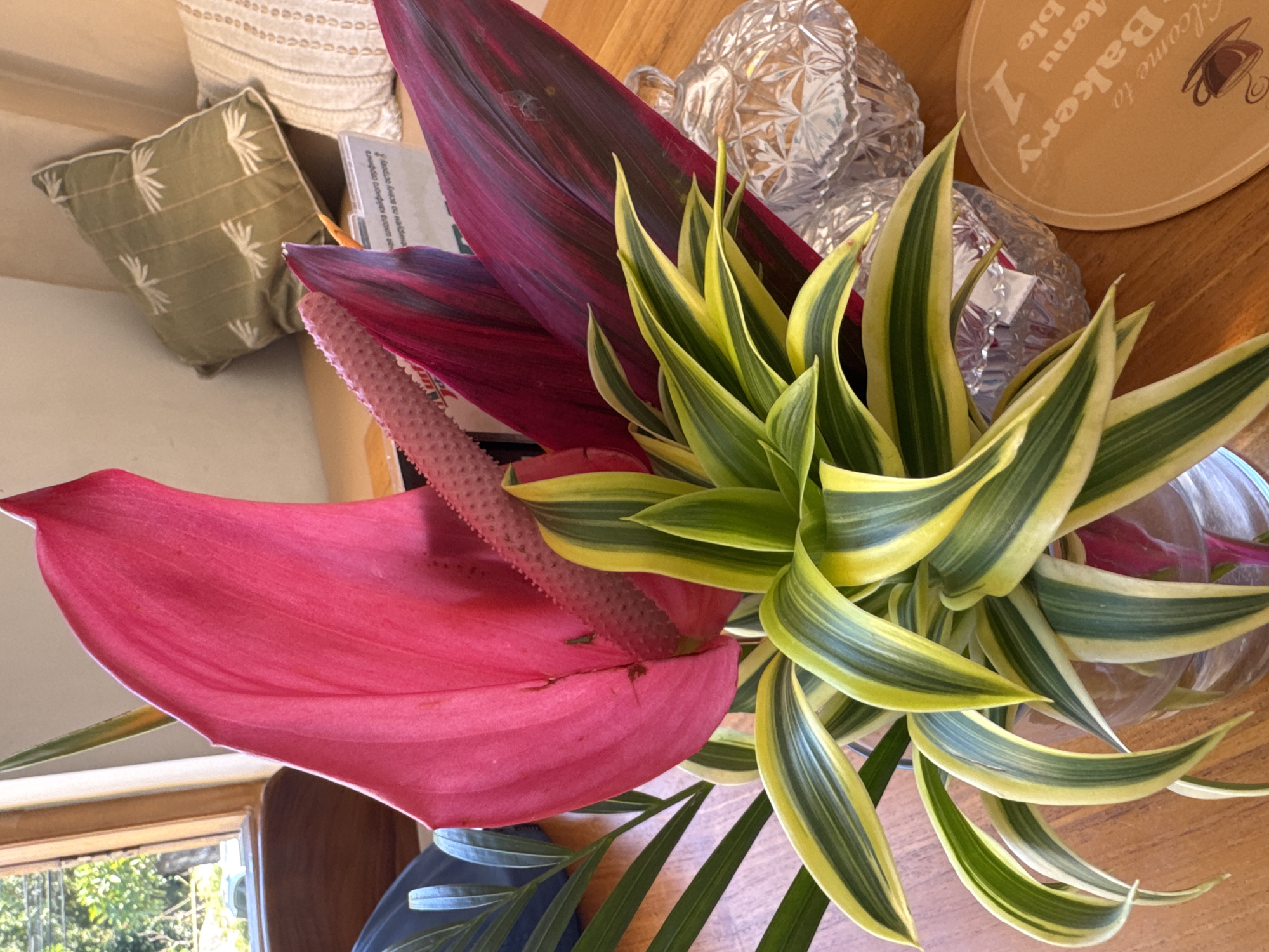 The image is a close-up, high-angle shot of a floral arrangement placed on a wooden surface. Prominently featured is a large, bright pink anthurium flower with a textured spadix. Behind the anthurium is a deep burgundy leaf, possibly a ti leaf (Cordyline fruticosa). To the right, there is a plant with yellow and green variegated leaves, likely a cultivar of Dracaena. The arrangement is situated next to several objects, including a clear, cut-glass container, a tan-colored coaster or menu with the words "Welcome to Bakery" printed on it, and a book. In the background, partially visible, is a throw pillow. The scene is likely indoors near a window, as suggested by the natural light. The city and Kuta Selatan, Indonesia were not mentioned and not possible to extract from the image.