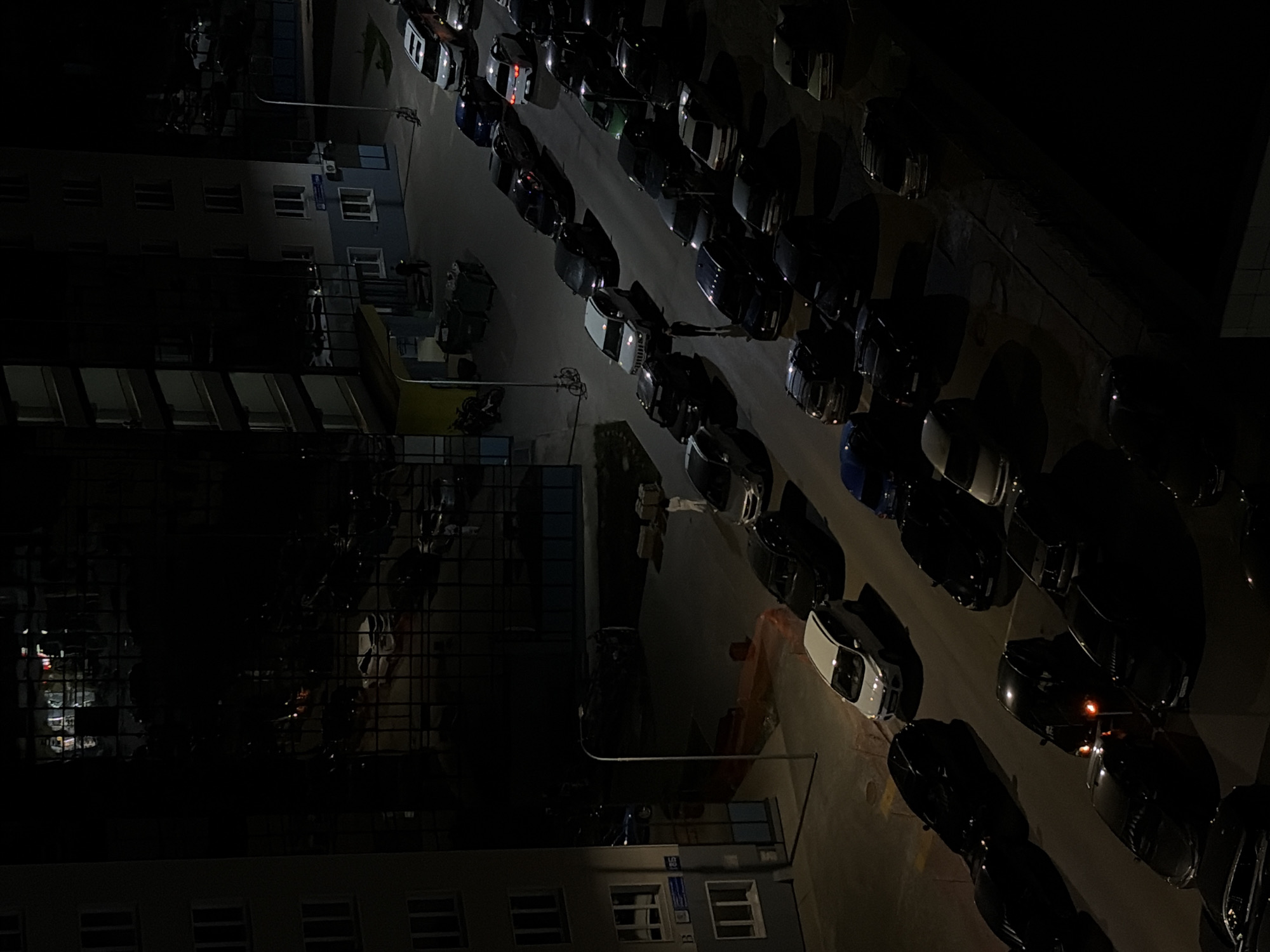 The image shows an aerial night view of a street in Naberezhnye Chelny, Russia, with multiple parked cars lining both sides. The street is illuminated by streetlights. A tall building is on the left side of the frame. The image appears to have been taken from a high vantage point, possibly from a building across the street. There are no people clearly visible.