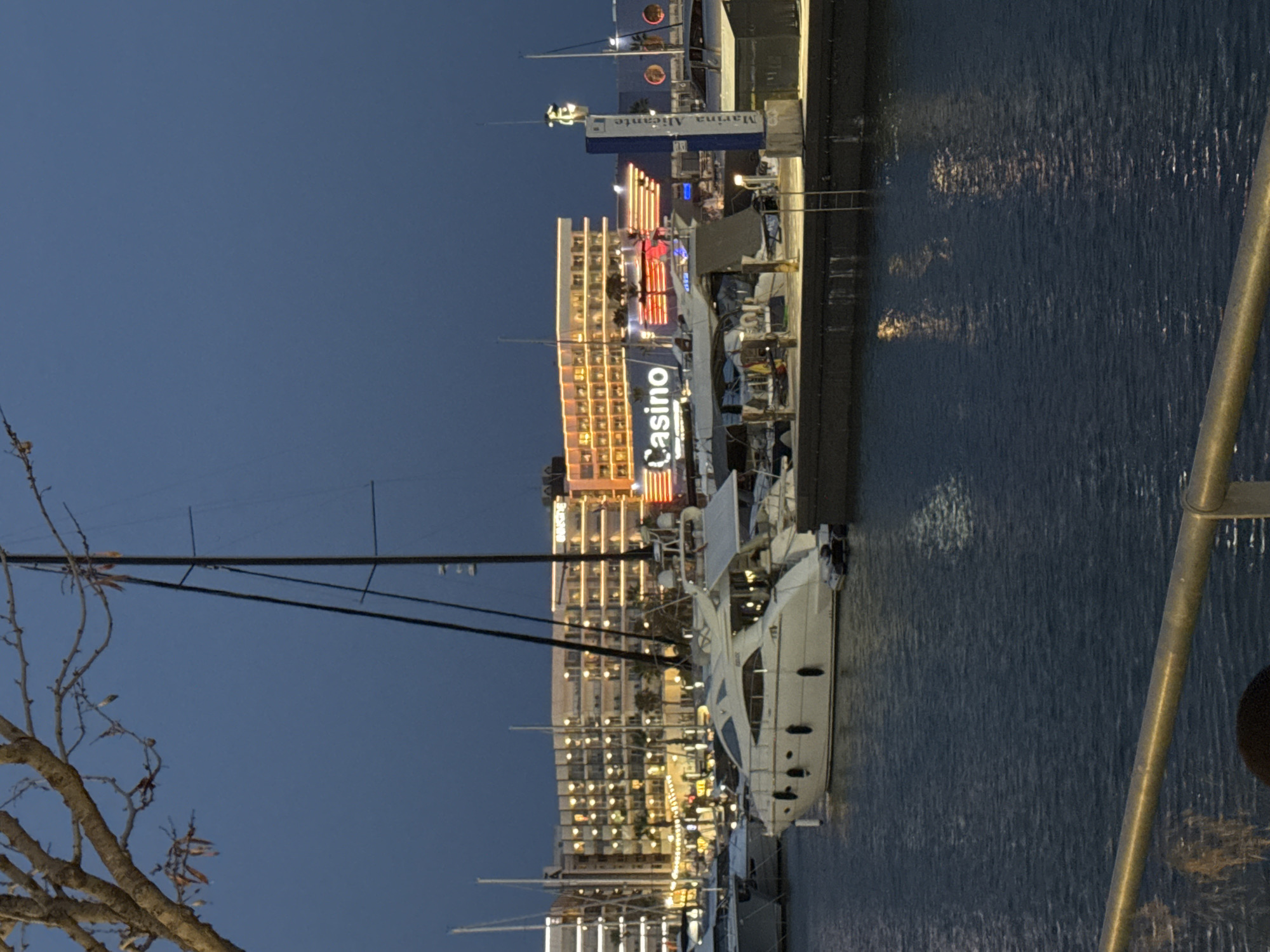 The image captures a waterfront scene at dusk in Alicante, Spain. In the foreground, a section of a metal railing is visible, with the dark water of the marina reflecting the lights of the buildings. A large white yacht is moored in the middle ground, with other boats and masts also present. In the background, a multi-story building with numerous illuminated windows and a prominent "Casino" sign is a focal point. Adjacent to the casino, a sign reads "Marina Alicante". The sky is a deep blue, indicating the late hour. The scene is well-lit by artificial lights from the buildings and boats.