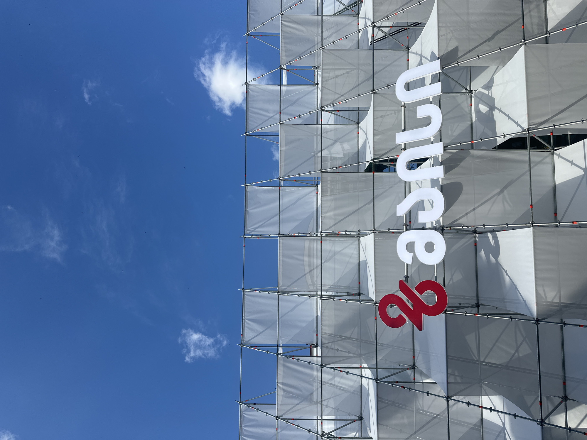 A low-angle shot displays a building facade obscured by a scaffold covered in white fabric panels, a clear blue sky with light clouds filling the background. The scaffold is constructed of metal poles with orange connectors. The word "Suu23" is vertically displayed in large, white and red letters against the scaffold.