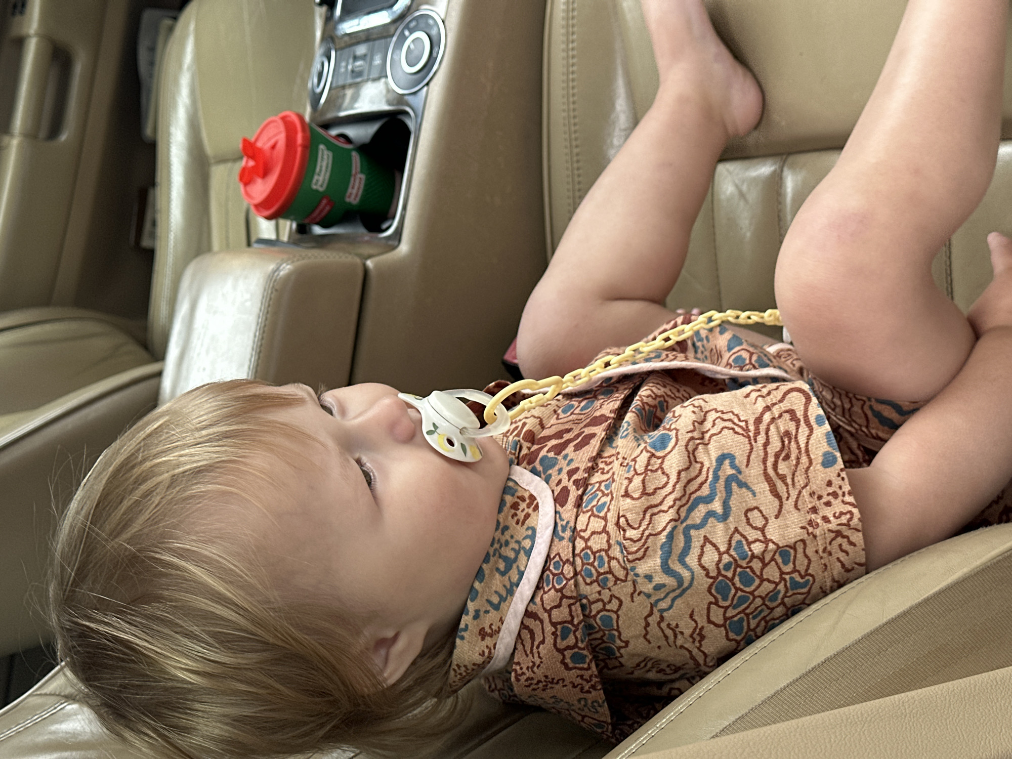 The image shows a toddler with blonde hair sitting in a car, wearing a patterned shirt and shorts. The toddler has a pacifier in their mouth. A red and green cup is visible in the car's center console. The car's interior appears to be made of leather. The image is a close-up shot, with the toddler's face and upper body being the main focus. The background is blurred, suggesting the car is in motion. There is no indication of the image being taken in Nizhny Novgorod, Russia, or any specific city.