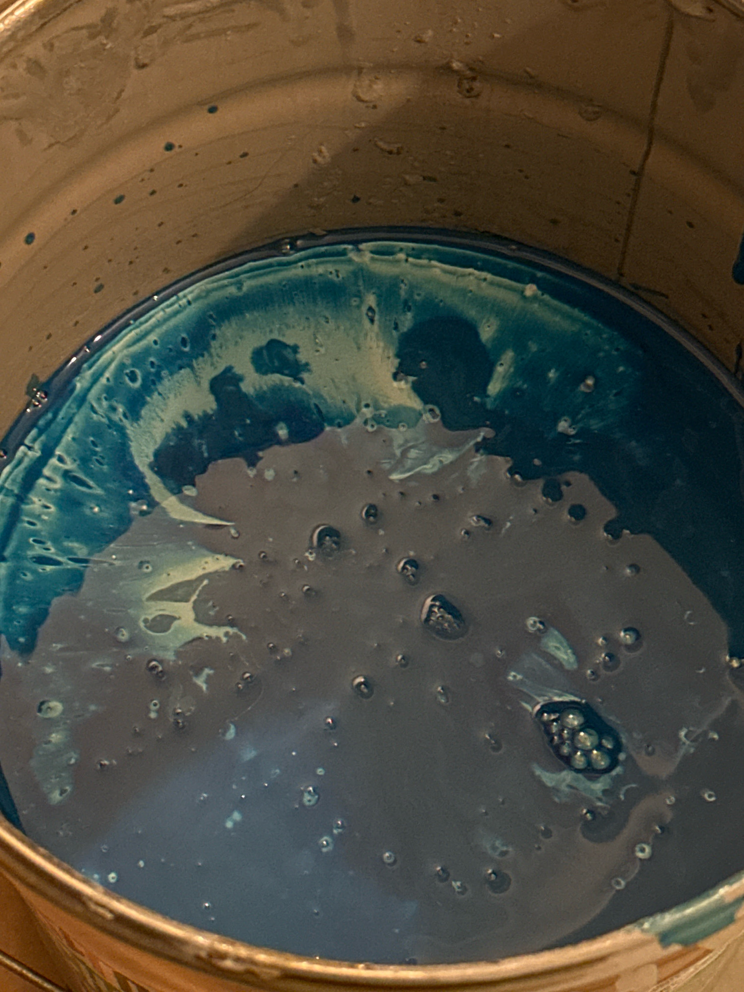 The image shows a close-up, overhead view of the inside of a metal bucket containing a dark blue liquid with swirls of lighter blue and white. There are numerous small bubbles on the surface of the liquid and some droplets on the interior wall of the bucket. The metal rim of the bucket is visible at the bottom and edges of the image, showing signs of wear and some dried residue. No people or specific locations within Nevinnomyssk, Russia are identifiable in this image.
