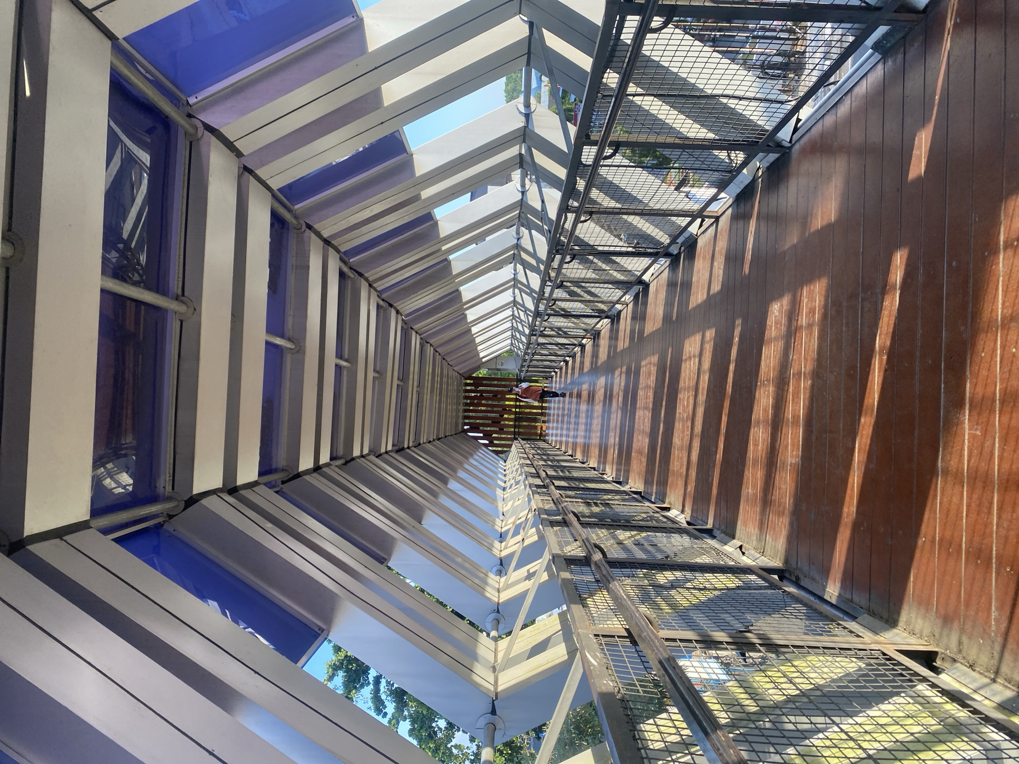 The image depicts an elevated walkway, likely a pedestrian bridge, in Banjarbaru, Indonesia. The walkway features a distinctive architectural design with a sloping roof supported by angled beams. The roof has sections of translucent blue panels interspersed with white or light-colored panels, allowing sunlight to filter through and cast patterned shadows on the wooden floor. Metal railings with a mesh infill line both sides of the walkway. A lone individual, wearing a reddish-brown top and dark pants, is walking away from the camera in the distance, towards the end of the bridge where a wooden structure is visible. The surroundings visible through the gaps in the roof and railings suggest an urban environment with trees and buildings under a clear blue sky. The overall impression is one of a modern, well-lit public space designed for pedestrian movement.
