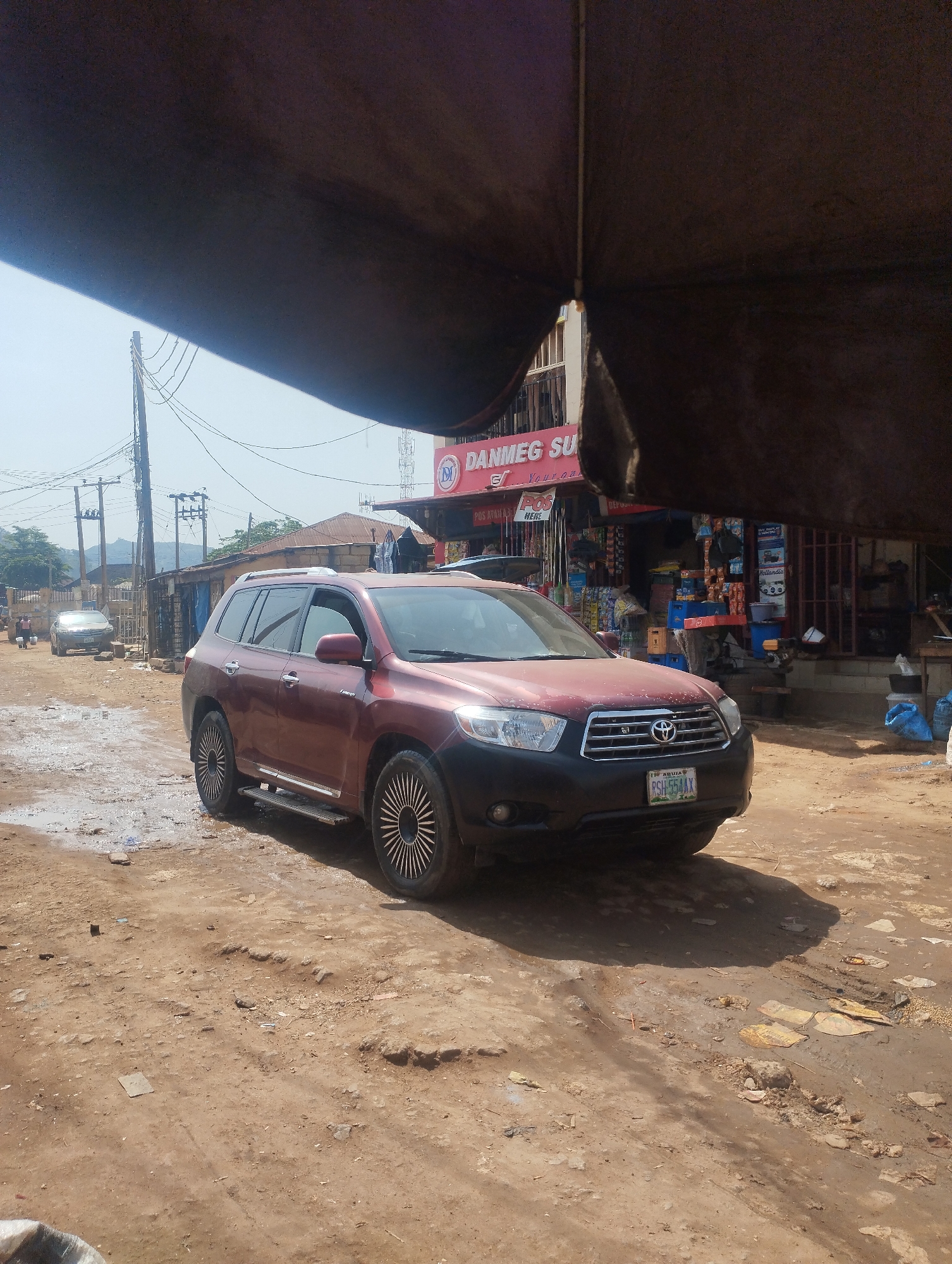 A maroon Toyota Highlander SUV is parked on a muddy, uneven dirt road in front of a small store with the sign "Danmeg Su" (partially obscured). The car has custom-looking black and silver wheels and a Nigerian license plate (RSH-554AX). The store appears to sell a variety of household goods. An awning partially obscures the top of the image. In the background, there are power lines and other buildings. The location appears to be Dutsen Alhaji, Nigeria based on the license plate and general scene. There are people visible in the distance near another parked car. The environment seems to be a busy commercial area, possibly a market street.