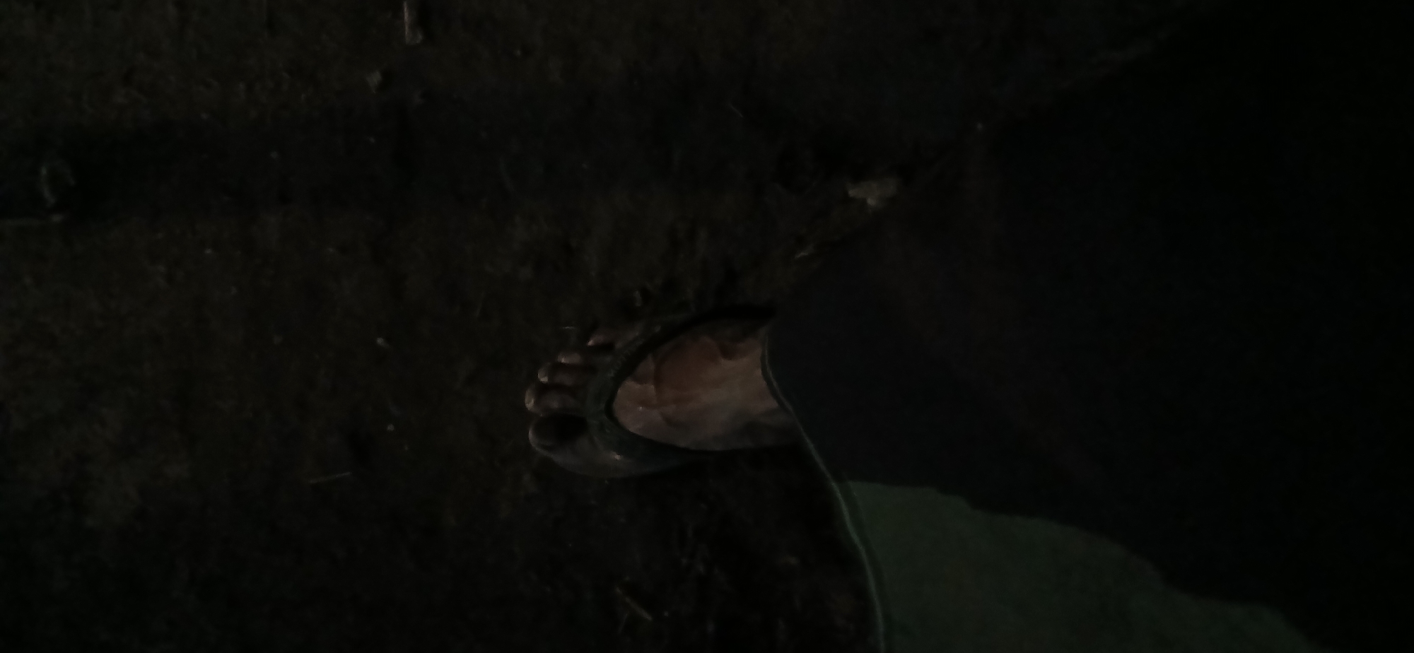 The image shows a close-up, low-angle view of a person's left foot wearing a dark sandal, resting on dark, textured ground. The foot appears to be bare and has visible veins. A dark fabric, possibly clothing or a mat, is partially visible in the foreground, along with a sliver of a greenish fabric. The lighting is dim, creating deep shadows and making details of the environment difficult to discern. The scene suggests a ground-level perspective, possibly in an outdoor setting with loose soil or dirt. The location is Kademangan Kulon, Indonesia.