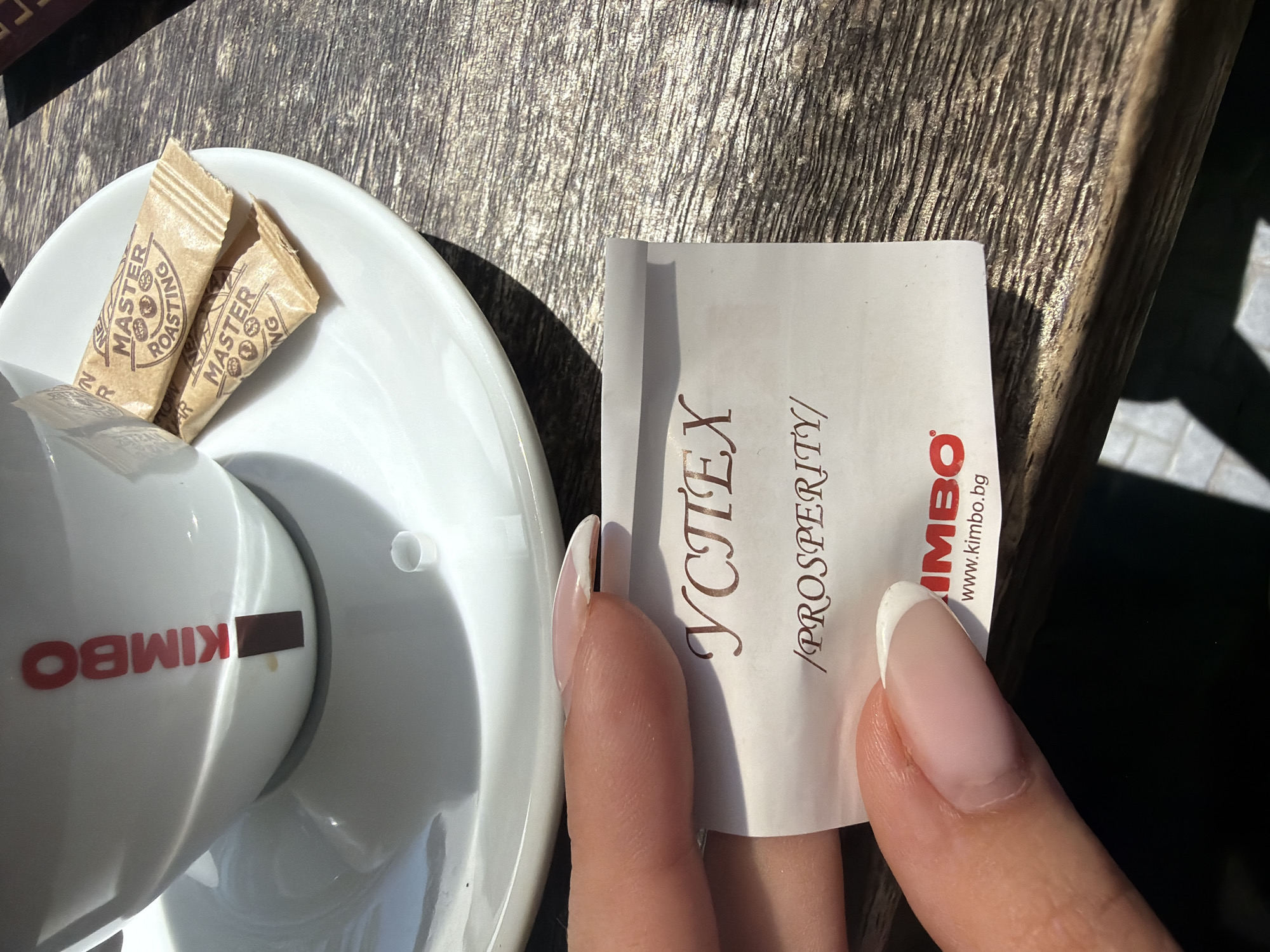 The image shows a close-up of a woman's hand holding a small white paper slip with text in both Cyrillic and Latin scripts. The slip is placed over a saucer with a white coffee cup and two sugar packets. The cup has the "KIMBO" logo in red. The sugar packets are branded "MASTER ROASTING". The paper slip has "УСПЕХ" and "/PROSPERITY/" printed on it, along with "KIMBO" and "www.kimbo.bg" at the bottom. The scene appears to be set on a wooden table, likely outdoors due to the sunlight and shadows. There is no specific indication of the city of Varna, Bulgaria within the image itself, only the website ".bg" which suggests a Bulgarian origin.