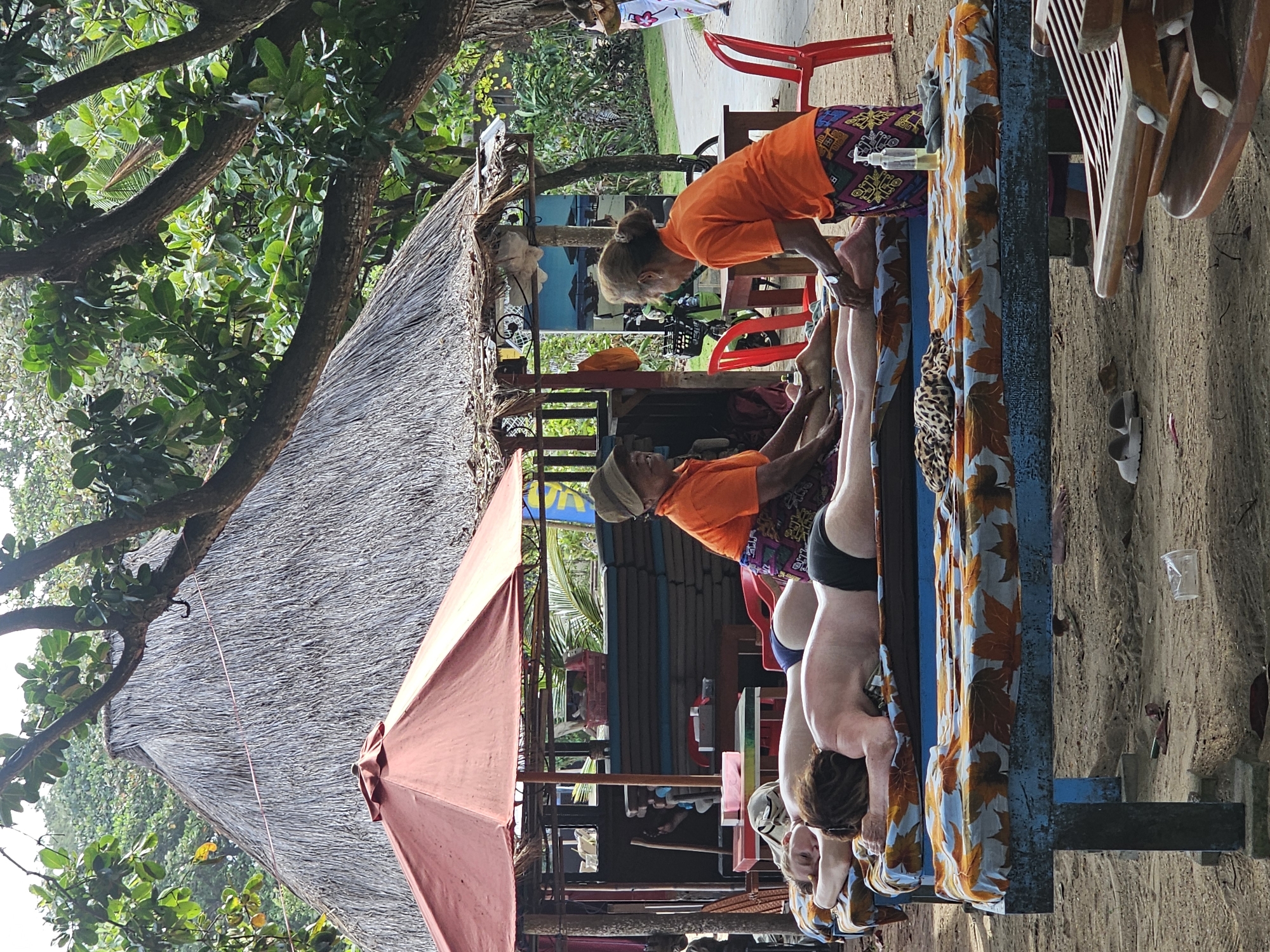 The image shows a scene at what appears to be an outdoor spa or massage area in Denpasar, Indonesia. Three people are lying face down on a massage table covered with a light blue patterned cloth. Two individuals are dressed in orange shirts and colorful patterned bottoms, likely massage therapists, bending over to work on the legs of the clients. There is a thatched roof overhead, partially covered by a pink umbrella. Red plastic chairs are nearby, along with other furniture and supplies used in a massage setting, like a bottle of oil or lotion. The ground appears to be sandy. The background features lush greenery and trees, typical of a tropical location.
