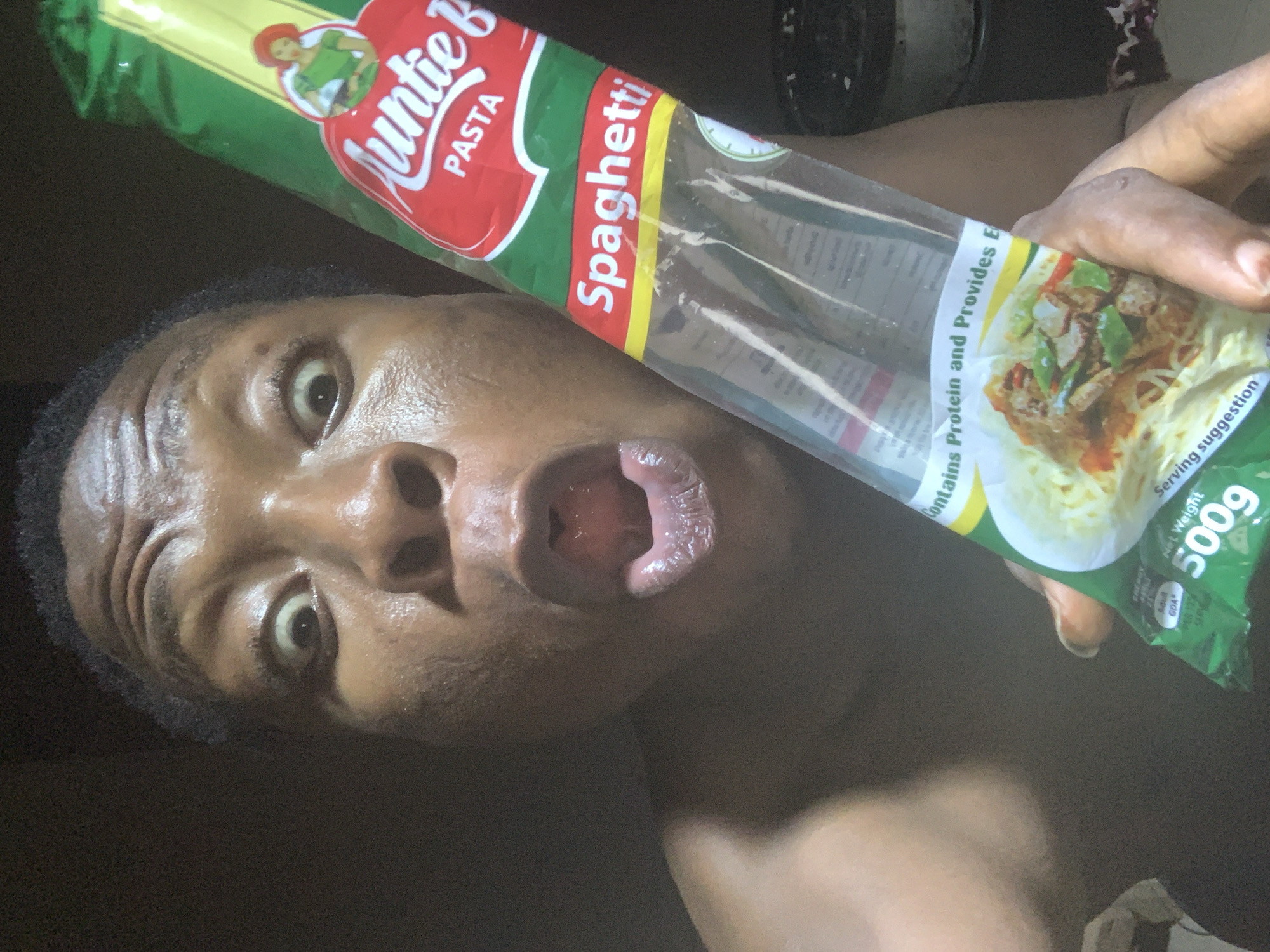 The image depicts a close-up of a person holding a packet of "Auntie B" brand spaghetti. The person's face is prominently featured, with wide eyes and an open mouth. The spaghetti packet is held in the person's right hand and displays the product name "Spaghetti" along with the brand logo and a serving suggestion image. The packet indicates a net weight of 500g. The background is dark and indistinct. No city or specific location details are discernible.