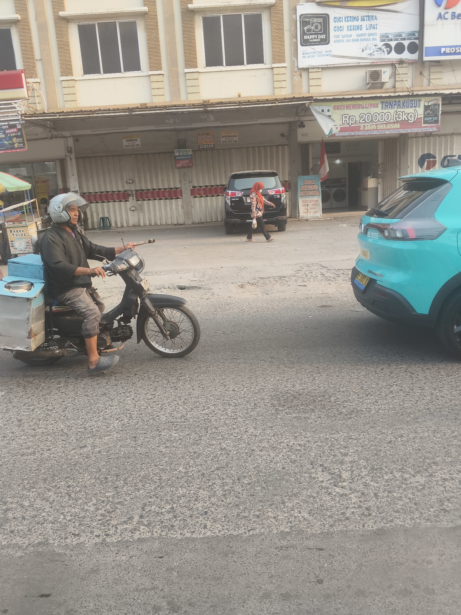 In a street scene in Bandar Lampung, Indonesia, an elderly man wearing a helmet, dark long-sleeved shirt, and grey pants rides a black motorcycle. He is wearing grey Crocs on his feet. The motorcycle has a light blue box attached to the rear, which appears to be for carrying goods. In the background, a black SUV is parked, and a woman in a light brown hijab and patterned clothing walks away from it. Adjacent to the SUV is a turquoise car. Buildings line the street, featuring storefronts with metal roll-up doors and signs for laundry services. One sign, in Indonesian, advertises "CUCI KERING SETRIKA" (Dry Cleaning Ironing) and mentions "RP. 20.000 (3kg)". The overall environment suggests a commercial area with businesses open for operation, and the lighting indicates it is daytime.