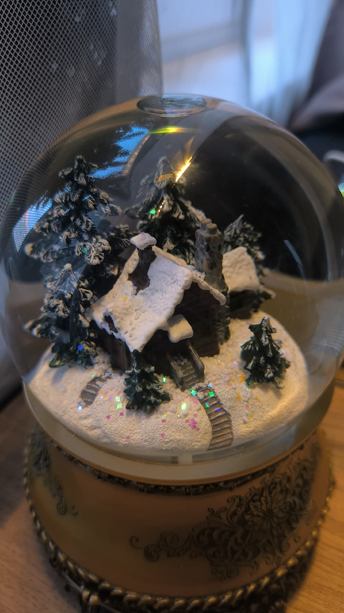The image shows a close-up view of a snow globe. The snow globe contains a miniature scene of a log cabin surrounded by snow-covered pine trees and pathways. Glittering specks are scattered throughout the scene, giving the impression of snow or confetti. The base of the snow globe is ornate, with a metallic gold finish and decorative detailing. The snow globe is positioned on a wooden surface. The background is blurred, but appears to include a grey mesh screen and a white curtain or fabric. The image is taken from a slightly elevated angle, looking down into the snow globe. There are no people present in the image, and no specific action is taking place other than the stillness of the scene within the snow globe. The location in the city of Kovrov, Russia, is not depicted or suggested by any elements in the image.