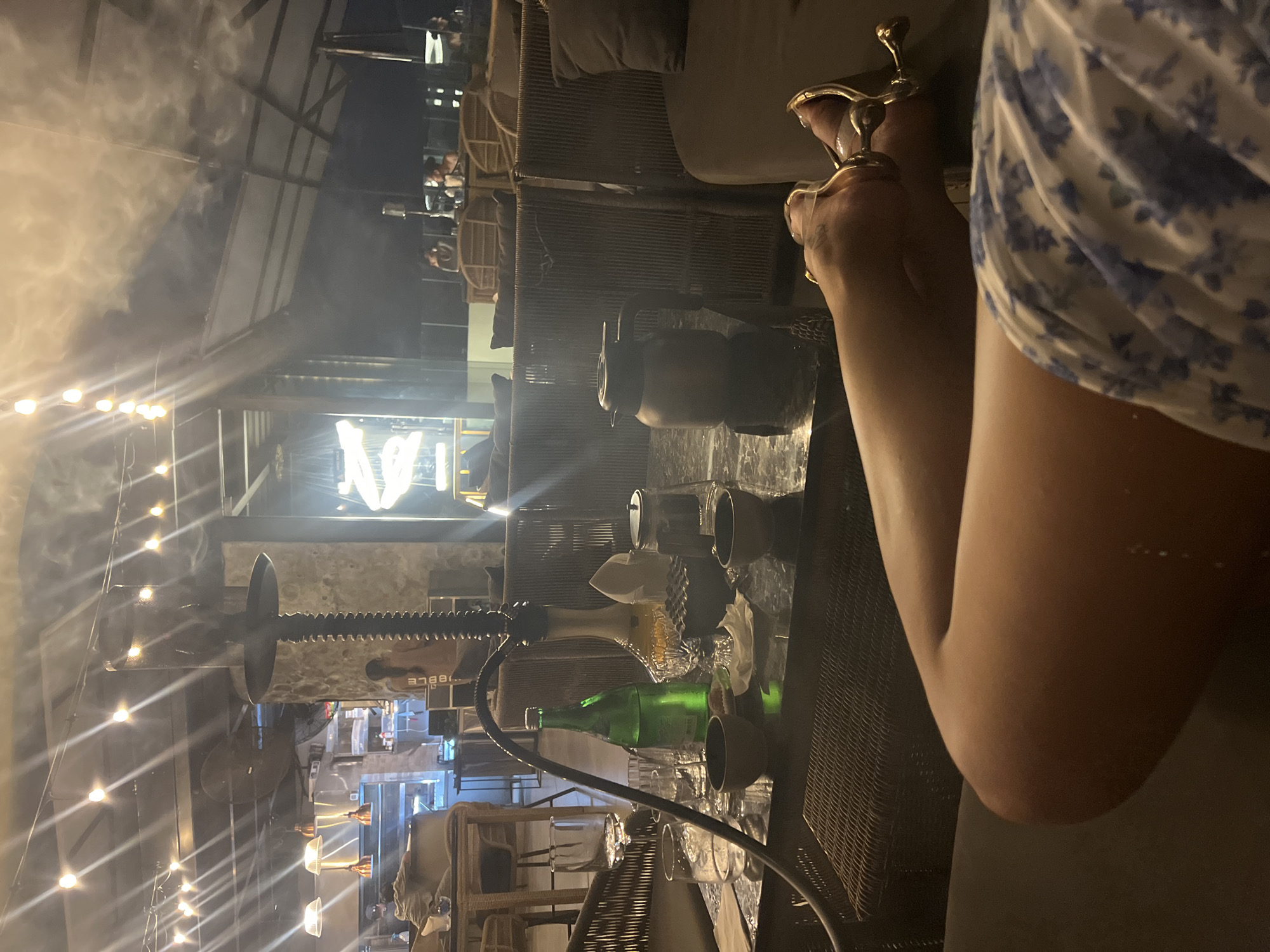The image portrays a dimly lit lounge or shisha bar at night in North Kuta, Indonesia, characterized by a relaxed and social ambiance. In the foreground on the right, a person is seated, their right arm and leg visible. They wear a light-colored dress with a blue floral pattern and clear sandals with gold accents.
A prominent shisha pipe with a black hose sits on a dark, woven table in the mid-ground. Also on the table are a green bottle, a decorative glass decanter, various glasses, and cups, suggesting beverages are being served. The air is hazy with smoke, likely from shisha.
The background reveals more of the establishment, featuring seating areas with woven rattan furniture and grey cushions. String lights draped across the ceiling emit a warm glow, illuminating the space. Other patrons are faintly visible in the distance, socializing. A large, bright sign displaying the letter "N" is clearly seen in the far background, with what appears to be "BBLE" faintly visible below a large, screw-like decorative object mounted on a wall. The architecture includes industrial-style beams and corrugated roofing elements. The overall atmosphere is intimate and convivial, typical of an evening gathering spot.