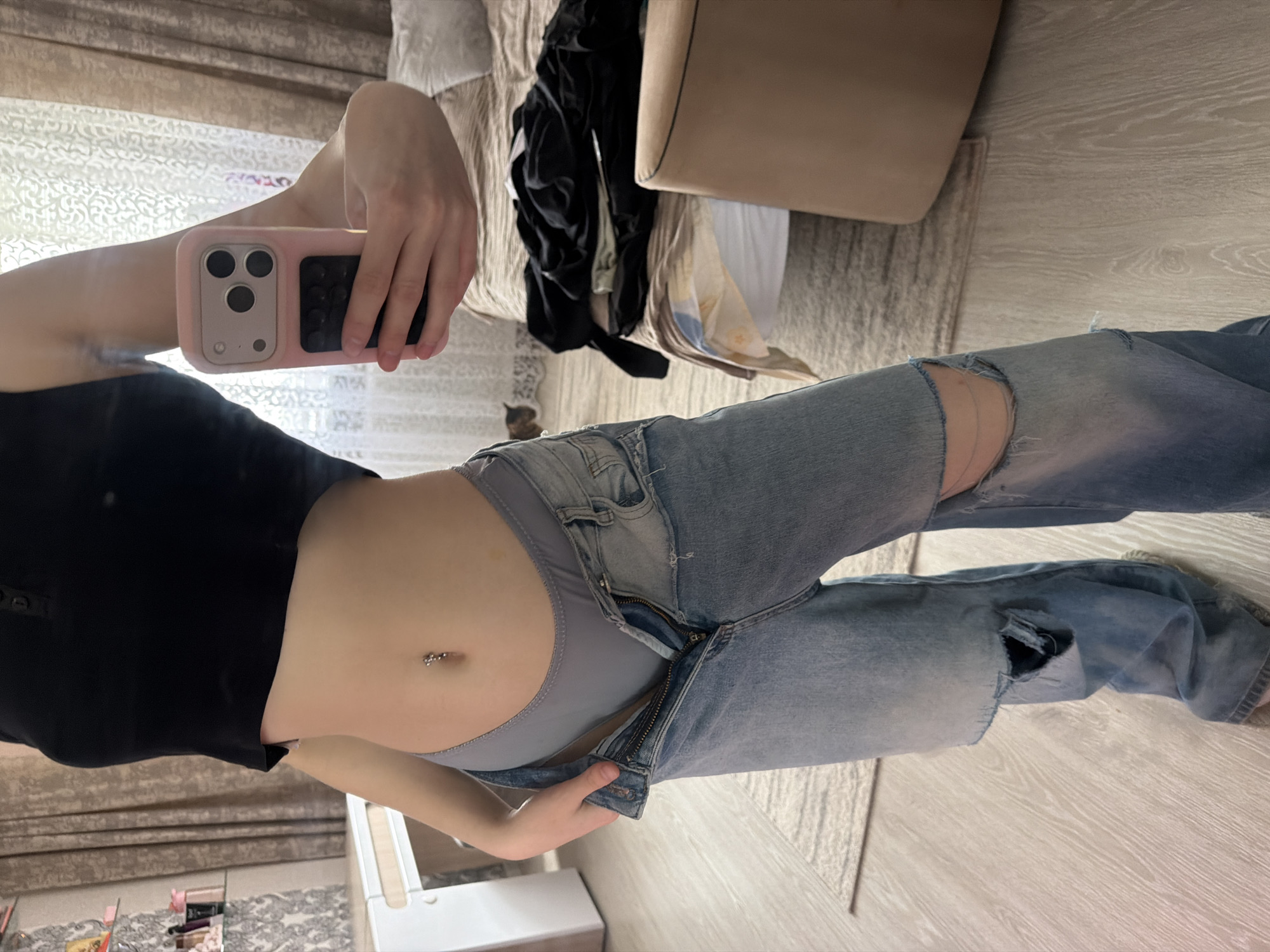 A person is seen taking a mirror selfie indoors, focusing on their torso and legs. The image is rotated 90 degrees clockwise. The person is wearing a black short-sleeved crop top and light blue ripped jeans, which are unzipped, revealing grey underwear. A belly button piercing with two small silver spheres is visible on their midriff.
In the upper left of the image (as rotated), the person holds a smartphone with a distinctive pink case that has a large camera cutout and a textured black panel on the back. Their right arm is slightly raised to hold the phone.
The background suggests a bedroom or dressing area. Behind the person, a bed or sofa is visible with various items of clothing, including black fabric, casually placed on it. To the right of the person, a light-colored upholstered chair or sofa armrest is partially visible. The floor is light-colored wood, and a patterned rug or carpet is also present. A small, brown cat or kitten can be faintly seen in the background between the person's leg and the bed. The lighting suggests it is daytime. There is no visible text in the image. The location context provided is Noviy, Russia.