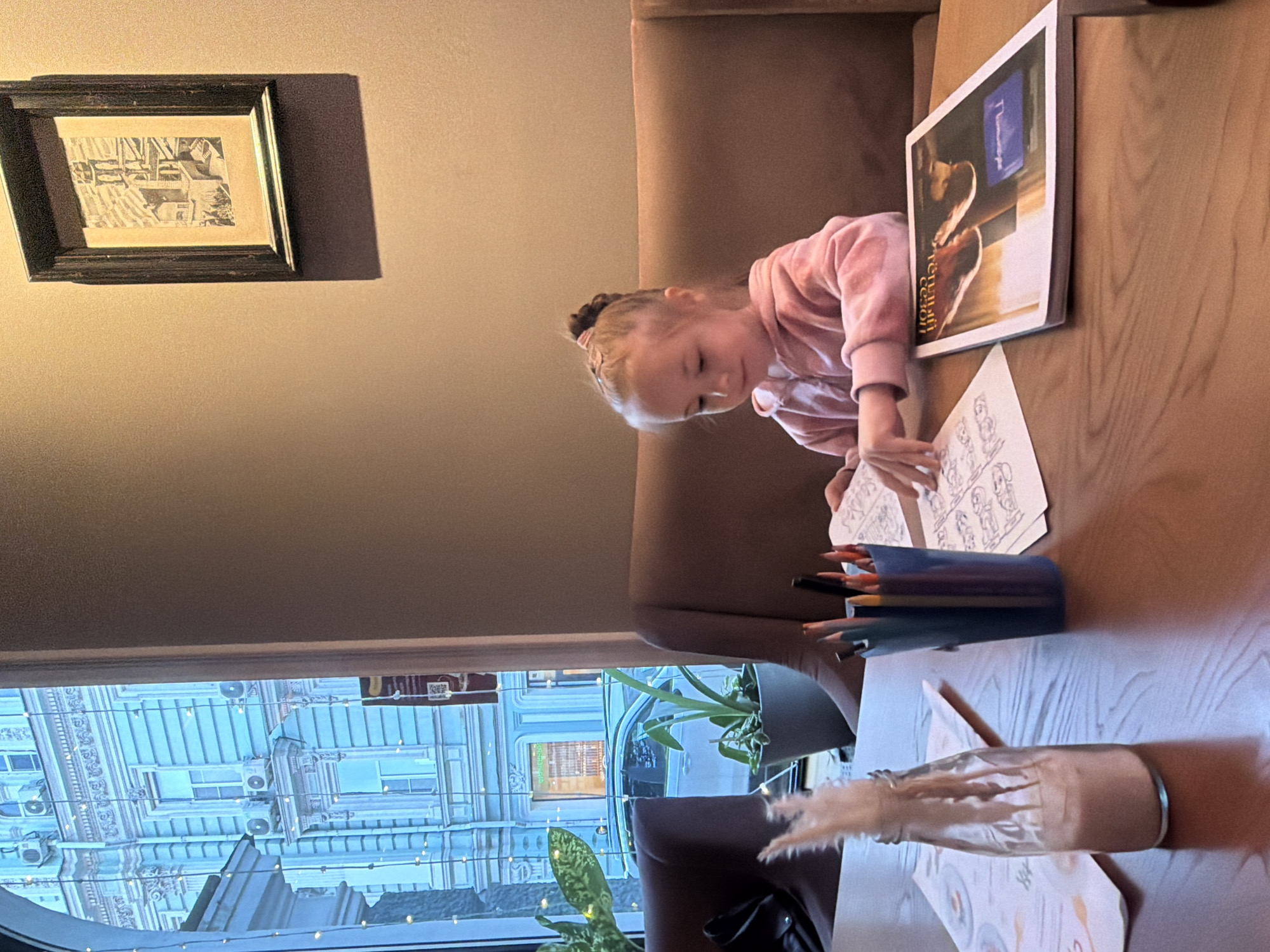A young girl is seated at a light-colored wooden table, intently coloring in a book. She has light-colored hair tied back and is wearing a pink hooded sweatshirt. Her hands hold a coloring utensil as she focuses on the outline drawings on the page in front of her.
The scene appears to be set indoors, likely in a cafe or restaurant, given the upholstered brown bench seating and the nature of the table. A container of colored pencils stands nearby on the table. To the girl's right, a magazine or book with a cover depicting pastries is leaning against something, displaying Cyrillic text "Творческий сезон" (Creative Season) and possibly "Лимонад" (Lemonade). Further down the table, a clear glass bottle containing a light brown liquid and some decorative feathery items is present, resting on another piece of paper with drawings, possibly a placemat or menu. A small potted plant adds greenery to the setting.
Behind the girl and above her, a plain light brown wall features a framed, monochrome picture depicting architectural structures. Through a large window to the left, an ornate urban building with multiple windows, air conditioning units, and decorative string lights is visible. The bright, diffuse light outside suggests it is daytime, possibly overcast or late afternoon. The girl appears calm and engaged in her activity.