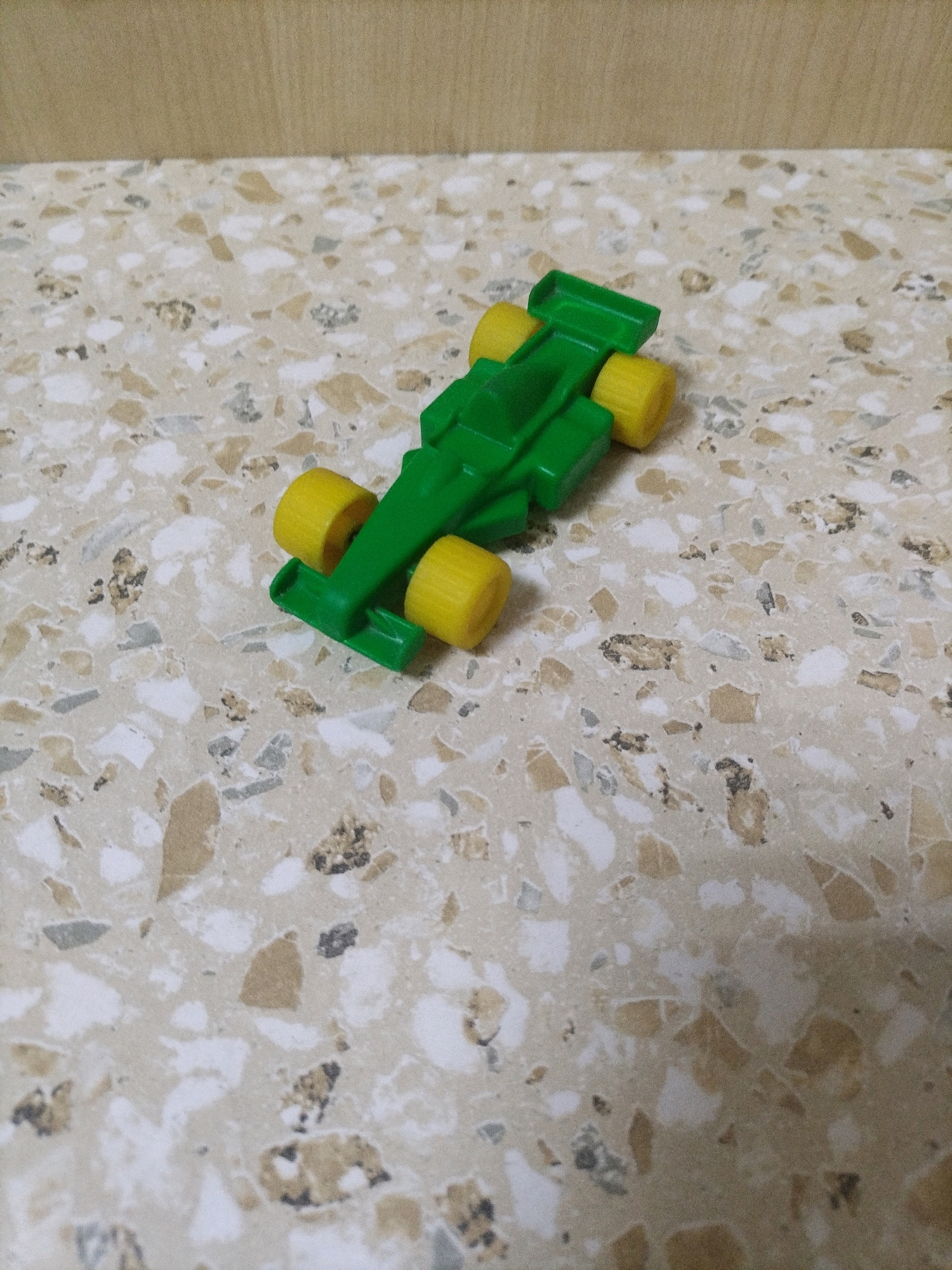 A green toy formula one race car with yellow wheels sits on a speckled beige and white countertop. The car is positioned at an angle with the front of the car facing the bottom left corner of the image. The background is a wooden wall. The image was taken in Hrodna, Belarus.