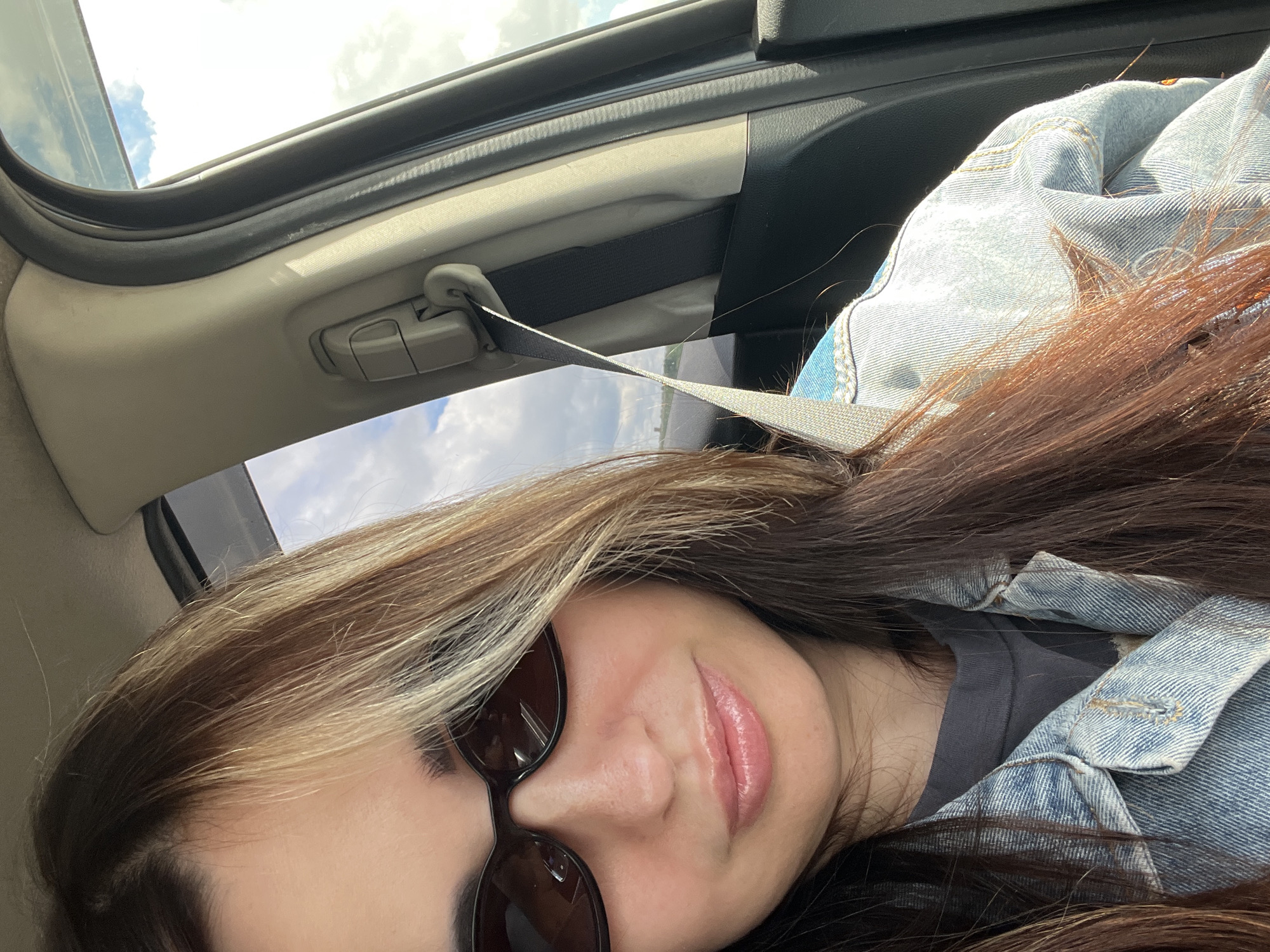A woman with long, dark brown hair with blonde highlights is sitting in the passenger seat of a car, wearing sunglasses and a denim jacket. She is looking towards the camera with a slight smile. The car's interior is visible, including a seatbelt and the window showing a cloudy sky. The background is out of focus, but appears to be a landscape. The image is taken from a selfie perspective.