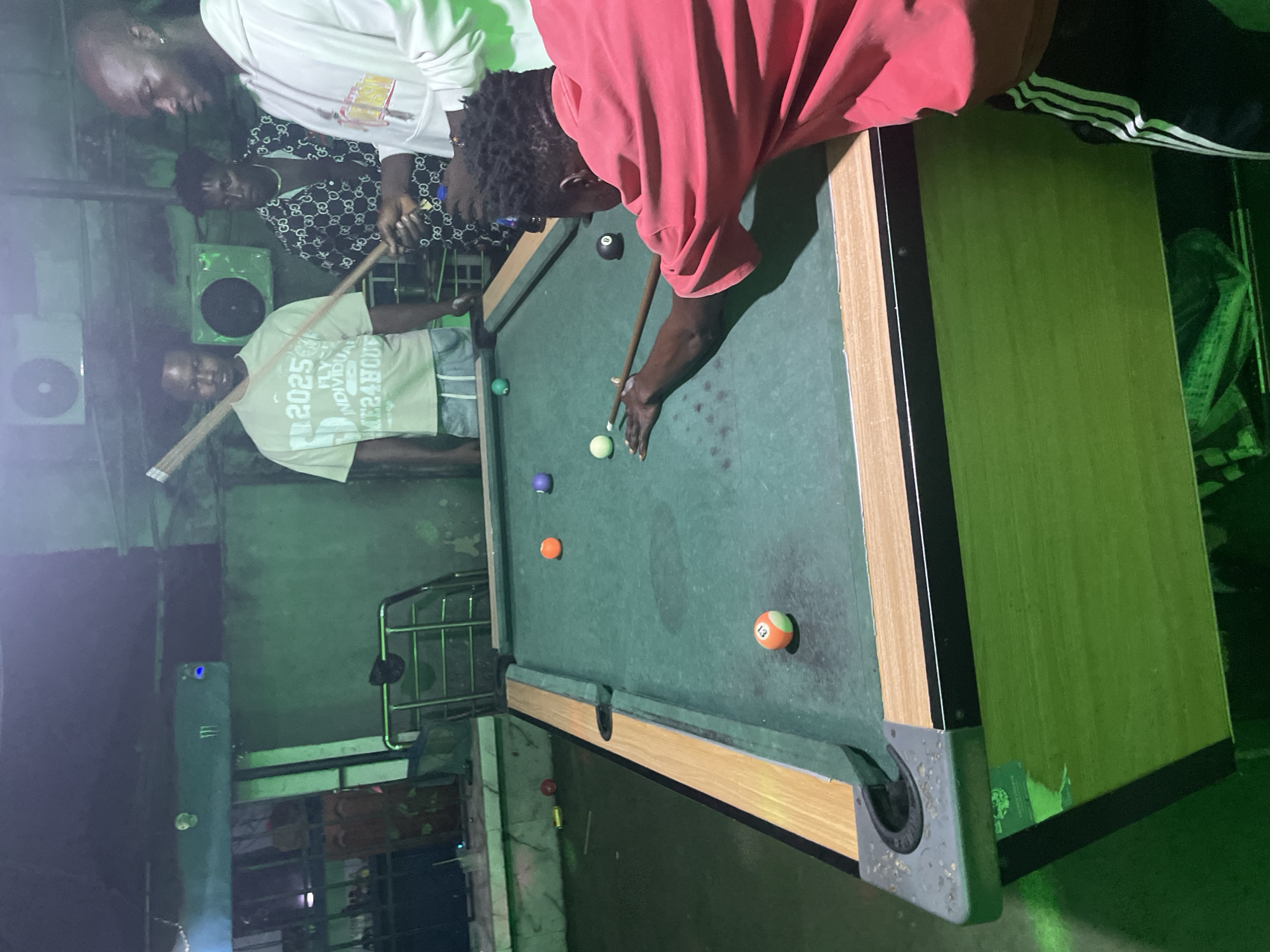 The image depicts a pool table in what appears to be a dimly lit, possibly outdoor or semi-outdoor space in Ado, Nigeria. A man in a pink shirt is taking a shot, with a pool cue touching the cue ball. Other people are watching, including a man in a white shirt with "2025" printed on it. An air conditioning unit is mounted on the wall in the background. The pool table has several balls on it, including an 8-ball and a striped ball. The overall environment appears to be casual and recreational.