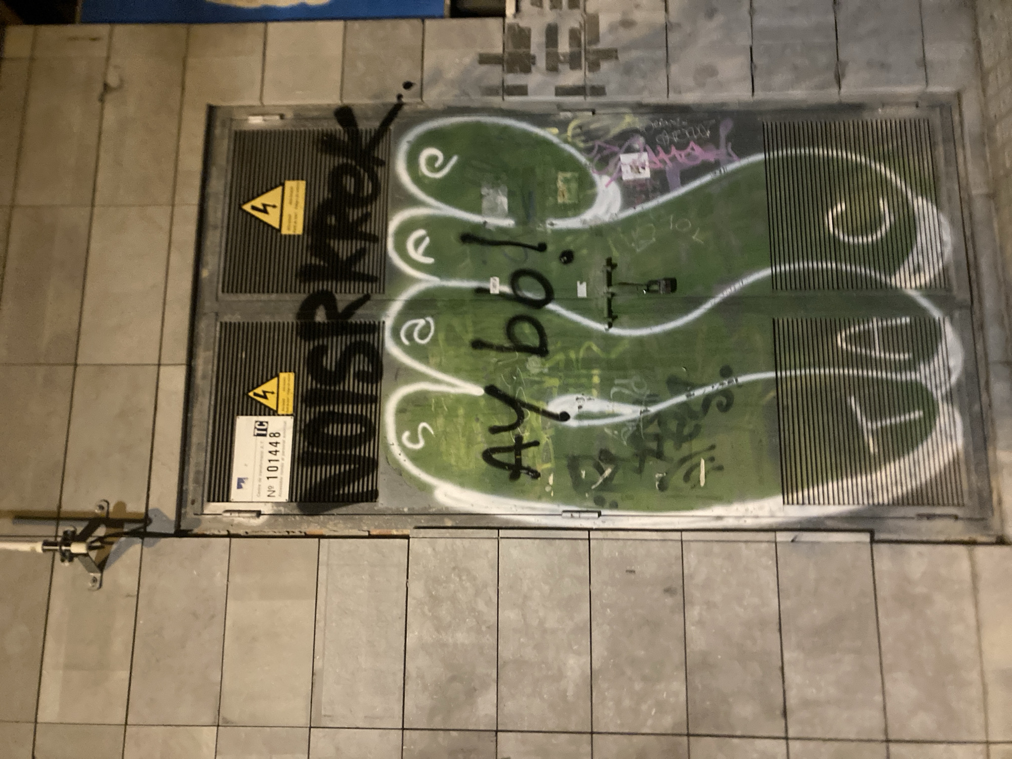The image is a medium-shot, eye-level, outdoor photograph of a metal utility cabinet covered in graffiti. The cabinet is located on a wall made of large, grey, rectangular tiles, suggesting an urban environment. The cabinet itself is metallic grey with a ribbed texture on the lower half of the doors. There are two prominent warning signs on the upper portion of the doors: one displaying a blue "TC" logo and a number "101448", and another with a yellow triangle and a black lightning bolt, indicating high voltage.
The graffiti on the cabinet is extensive and colorful. The most prominent graffiti is in black and white. A large black script reads "NOISRkREK" across the top of the doors. Below this, a large green shape outlined in white resembles an abstract figure or hand with three bulbous protrusions. Within this green shape, in black, are the words "AY bbl!". White graffiti also outlines the letters "T A C" on the lower section of the doors, appearing through the ribbed metal. There are also smaller tags and scribbles in various colors, including pink and yellow, scattered across the surface of the cabinet and wall. A small padlock is visible on the green graffiti. The overall scene is dimly lit, suggesting it was taken at night or in a poorly lit area.