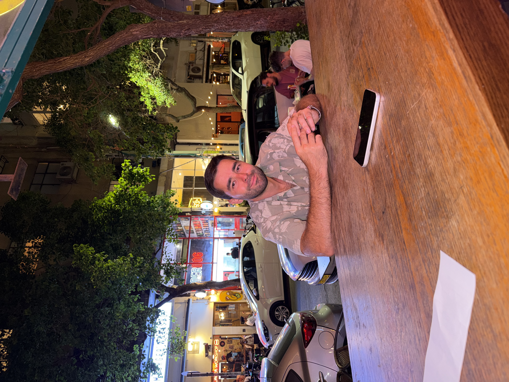 A man wearing a floral short-sleeved shirt is seated at a wooden table in an outdoor urban setting at night, with trees and illuminated buildings in the background. He is looking towards the camera with a slight smile. His hands are clasped on the table in front of him. A smartphone is lying on the table to his right, and a folded piece of white paper is to his left. In the background, cars are parked on the street, and people are visible at tables outside of establishments with neon signs. The scene appears to be in Tel Aviv, Israel, likely on a pedestrian-friendly street with restaurants and shops.