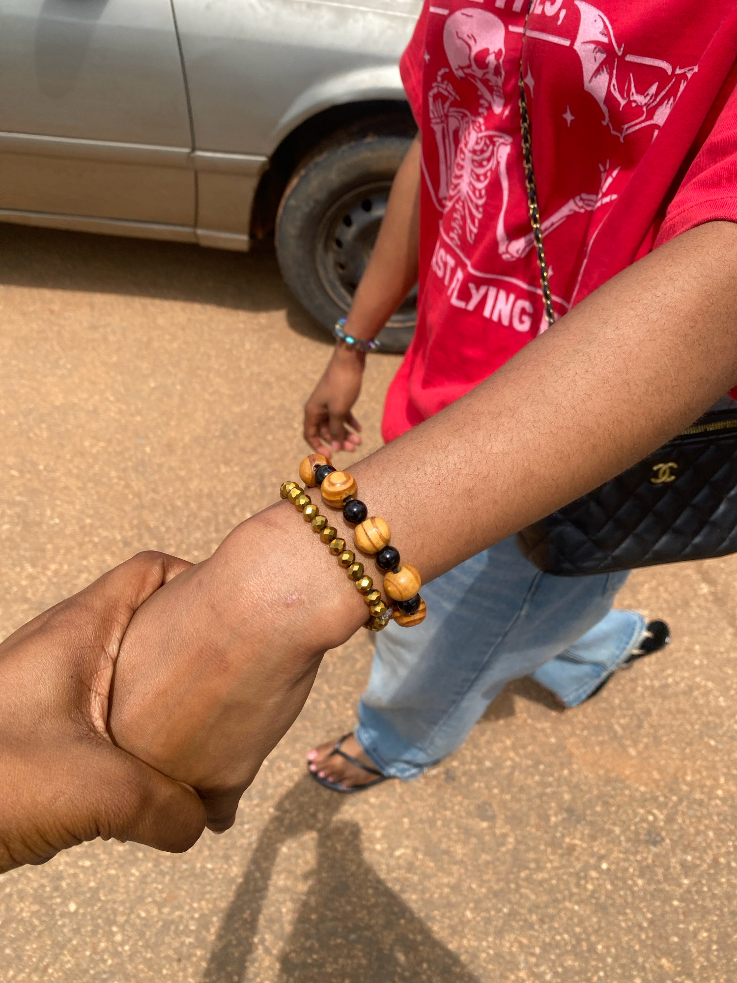 Two individuals' arms are clasped together, suggesting a moment of connection or support. One individual, wearing a red t-shirt with a skeleton graphic and the words "JUST LYING" printed on it, has a black quilted purse with a gold Chanel logo slung across their shoulder. They are also wearing blue jeans and flip-flops. The other individual's arm, adorned with beaded bracelets, is held by the first. The ground is a textured, reddish-brown surface, likely a dirt or paved road, and a portion of a silver car is visible in the background, including its wheel and fender. The scene appears to be outdoors, possibly in a public area within the city of Iworoko, Nigeria, given the dry, dusty environment and the style of the car.