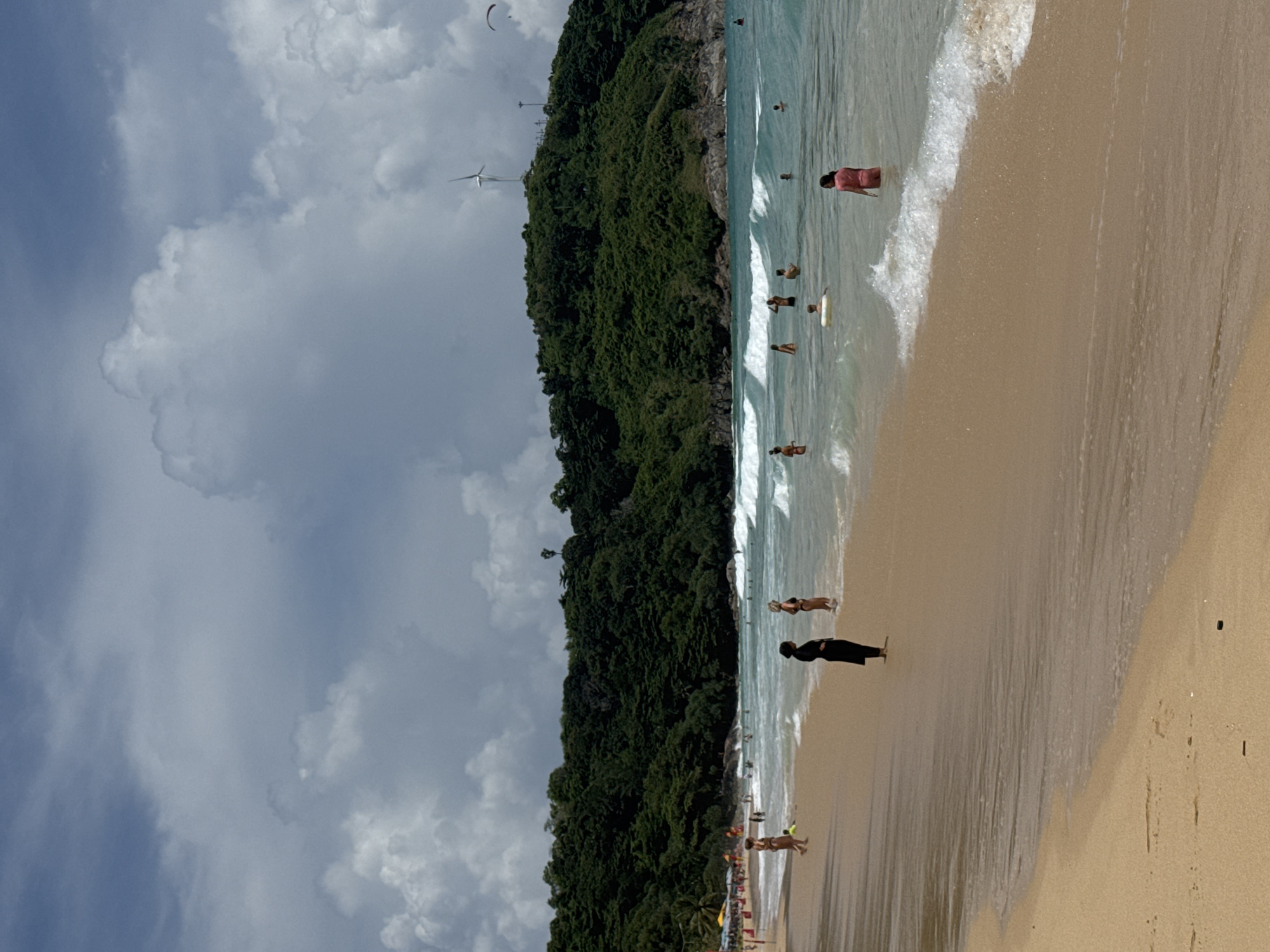 A wide, sandy beach in Rawai, Thailand, is populated with a moderate number of people enjoying the water and shoreline. The ocean features moderate waves, with white crests indicating active surf. A lush, green, tree-covered hill forms the backdrop to the beach. On top of the hill, a prominent wind turbine is visible, along with what appears to be a paraglider in the sky to the right. Several individuals are wading in the shallow water, while others stand on the wet sand near the shoreline. Some are dressed in swimwear, and one person is wearing a black cover-up. The sky is partly cloudy, with a mix of white and darker gray clouds. The sand on the beach is a light tan color, and the water is a clear turquoise.