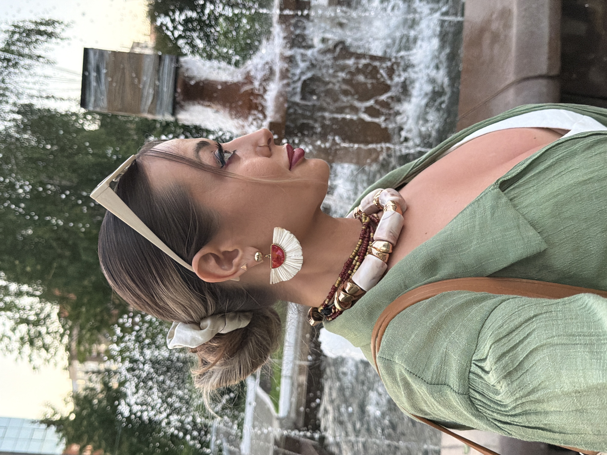 This image, reportedly taken in Krasnoyarsk, Russia, depicts a female individual in a right-side profile view, looking towards the right side of the frame. Her brown hair, with lighter highlights, is pulled back into a messy bun secured by a light, off-white fabric scrunchie. Light-colored sunglasses are pushed up on her head. She wears a small gold-toned triangular stud earring near the top of her right ear, and a larger, fan-shaped dangle earring below, featuring white fringe, a red semi-circular base, and a gold-toned stud. A subtle nose stud is visible on her right nostril. Her makeup includes dark eyeliner and dark red lipstick. She is wearing an olive green, textured, long-sleeved collared top, with a white garment visible underneath its plunging neckline. A brown strap, likely from a bag, crosses over her right shoulder. The background is heavily blurred (bokeh), showing a large water fountain with prominent spray and droplets, along with indistinct trees and structures in the distance, suggesting an outdoor setting during ambient light conditions, possibly late afternoon or early evening.