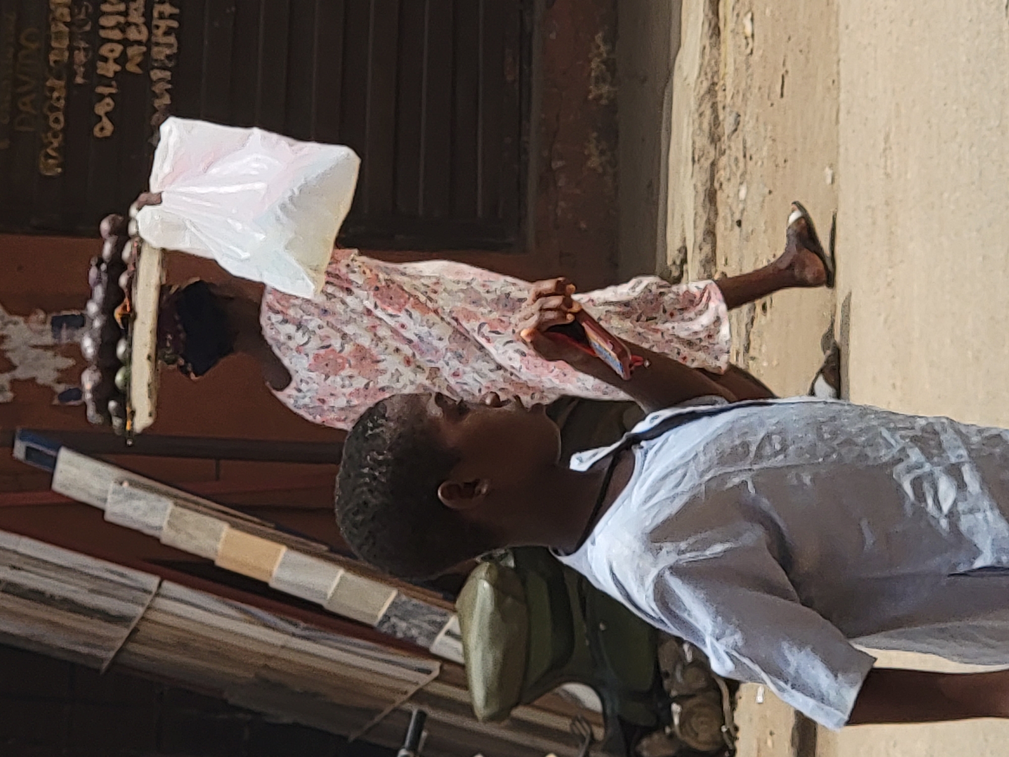 The original image is rotated 90 degrees counter-clockwise, depicting an outdoor street or market scene, likely in Maiburiji, Nigeria, under bright daylight.
In the foreground, a young boy with short, dark hair is shown in profile, wearing a light-colored short-sleeved collared shirt. He is looking upwards and to the left, observing something beyond the frame. Behind him, a large green object is partially visible, possibly a bag or part of a vehicle.
Further in the background, an adult figure, likely a woman, is visible. She is dressed in a floral-patterned dress and has a dark head covering. Her left hand holds a translucent white plastic bag filled with several small, dark, round items, possibly onions or similar produce. Her right hand is grasping a small, reddish-brown rectangular object, which could be a mobile phone or another small item. Her right foot, wearing a simple sandal, is visible against a light-colored, textured wall.
The ground appears to be a sandy or dusty path. Behind the adult figure, a dark-colored wall is visible, with handwritten text in white. Partially legible text includes "PAVDO" or "DAVDO", a partial phone number "08140199...", "MWE", "MINT", and what appears to be "LIIH7" below them. The strong illumination indicates a sunny day. The scene captures daily life with individuals moving along a street, possibly engaged in commerce.
