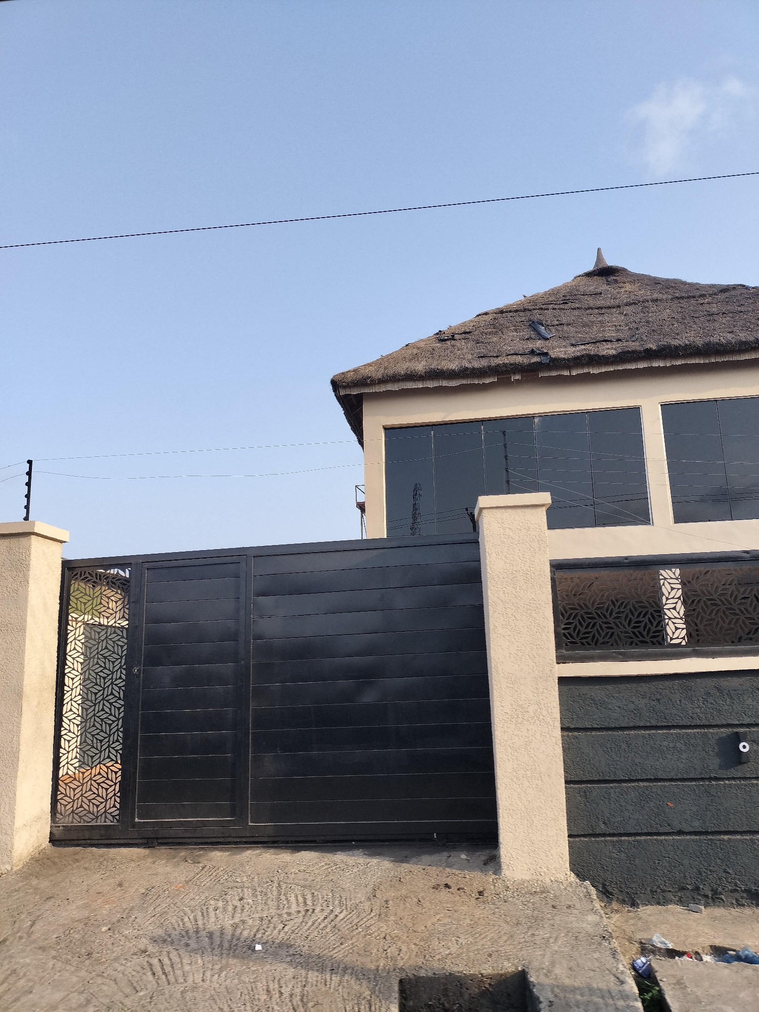 The image shows the exterior of a two-story building in Alaiyabiagba, Nigeria. The building has a thatched roof, cream-colored walls, and large, dark-tinted windows. A black metal gate with decorative geometric cutouts is positioned in front of the building, flanked by tan pillars. An electric fence runs along the top of the pillars. The ground in front of the gate is paved with concrete. Some litter is visible near the bottom right corner of the frame. The sky is blue and clear, and a power line is visible in the upper portion of the image. No people are visible.