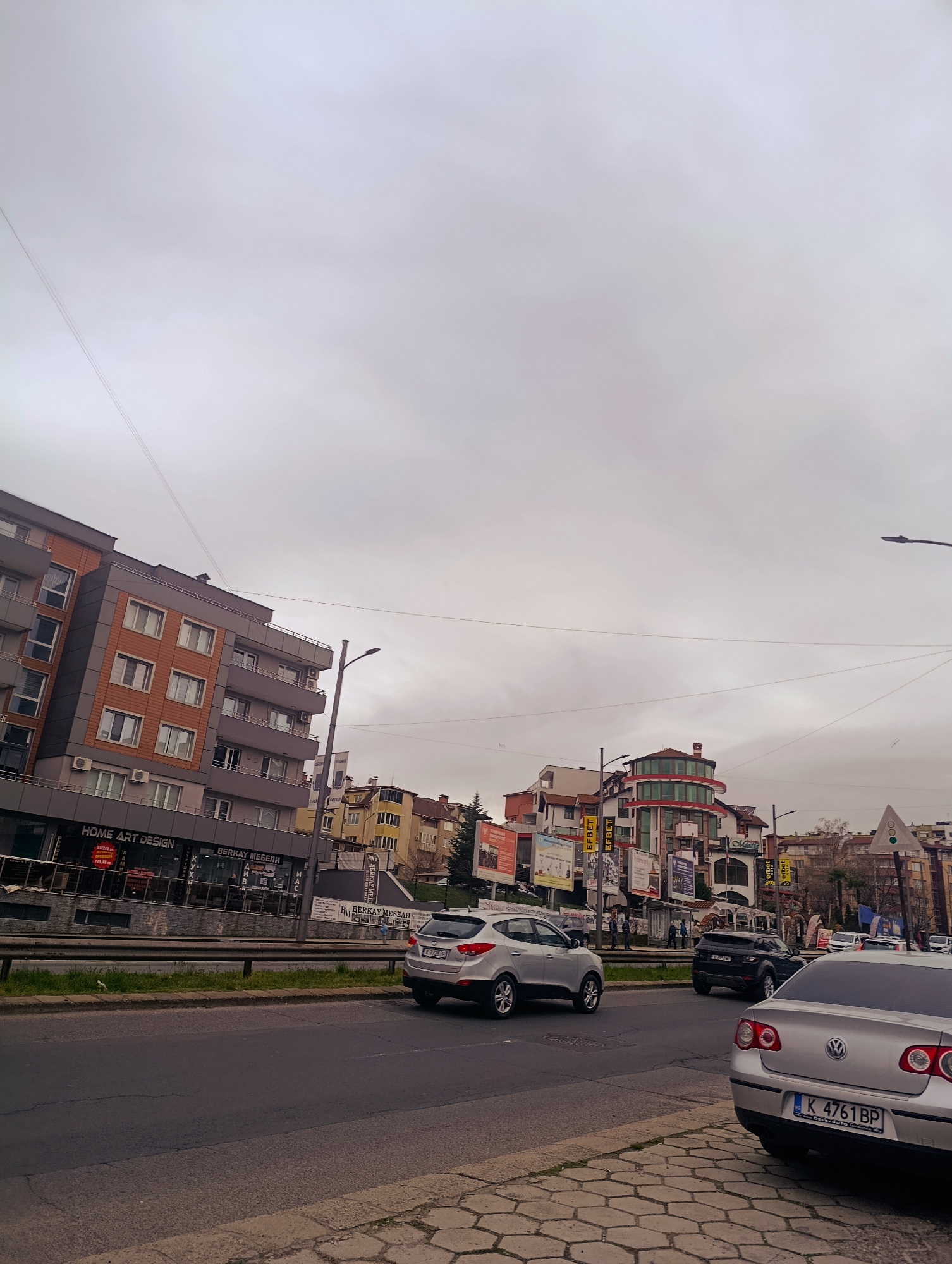 The image depicts an urban street scene in Kardzhali, Bulgaria on an overcast day. A gray building with "HOME ART DESIGN" and "BERKAY MEBELI" storefronts is visible on the left. Several other buildings are situated in the background. In the foreground, vehicles are on the road, including a silver Hyundai, a dark SUV, and a silver Volkswagen Passat with Bulgarian license plate "K 4761 BP". The sidewalk is paved with hexagonal stones. Signage and advertising posters are scattered throughout the scene, along with utility poles and wires overhead.