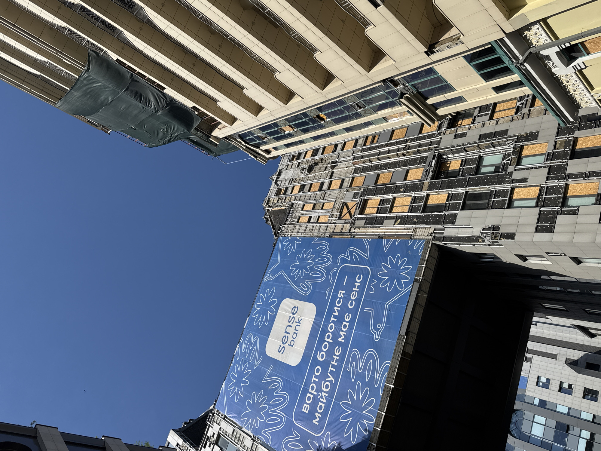 The image captures a view looking up at buildings in Kyiv, Ukraine. Some windows on a mid-rise building are boarded up with what appears to be plywood. A large blue banner for Sense Bank is displayed on the side of one building. The sky is clear and blue. There are some other buildings visible at the edges of the frame.