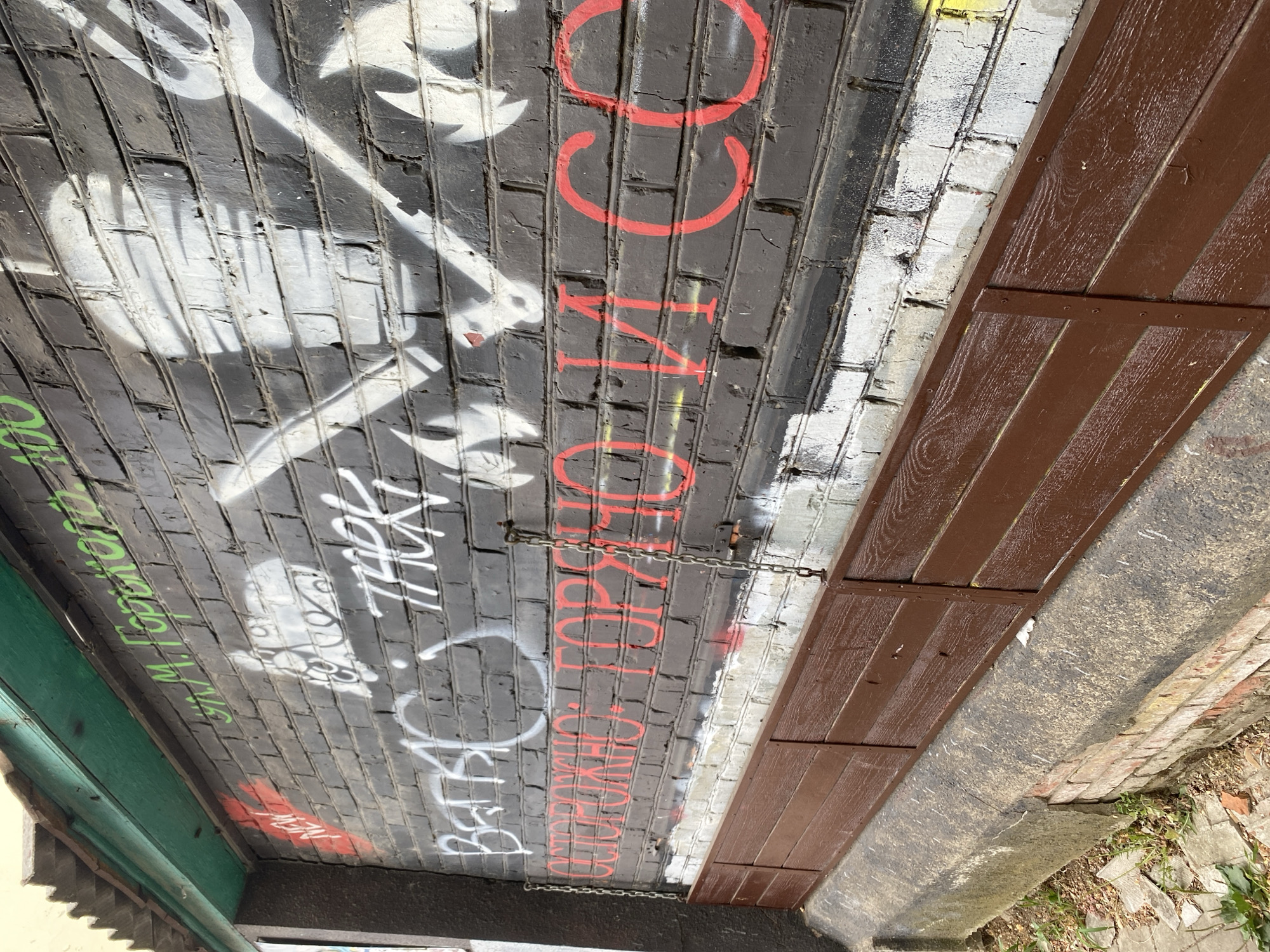 This image shows a brick wall covered in graffiti. The wall is painted black and has various spray-painted designs and text in white, green, and red. In the top left corner, green text reads "ул. М. Горького, 100" (M. Gorky Street, 100). To the left of this, red spray paint forms the word "NEW". Below this, white graffiti includes the word "ВЕРА" (Faith) and what appears to be a stylized drawing of a face. Prominently displayed in red, large lettering across the center of the wall is the phrase "ОСТОРОЖНО: ГОРЯЧО И СО" (Caution: Hot and Co). There are also white spray-painted silhouettes of figures, possibly ghosts or spirits, on the upper portion of the wall. A wooden structure, possibly a bench or planter box, is attached to the lower part of the wall. Metal chains hang from the wall to the wooden structure. The wall itself appears weathered and damaged in places, with peeling paint and visible brickwork. The area around the base of the wall shows some concrete and broken paving stones, with sparse green vegetation. The overall impression is that of an urban exterior with graffiti art.