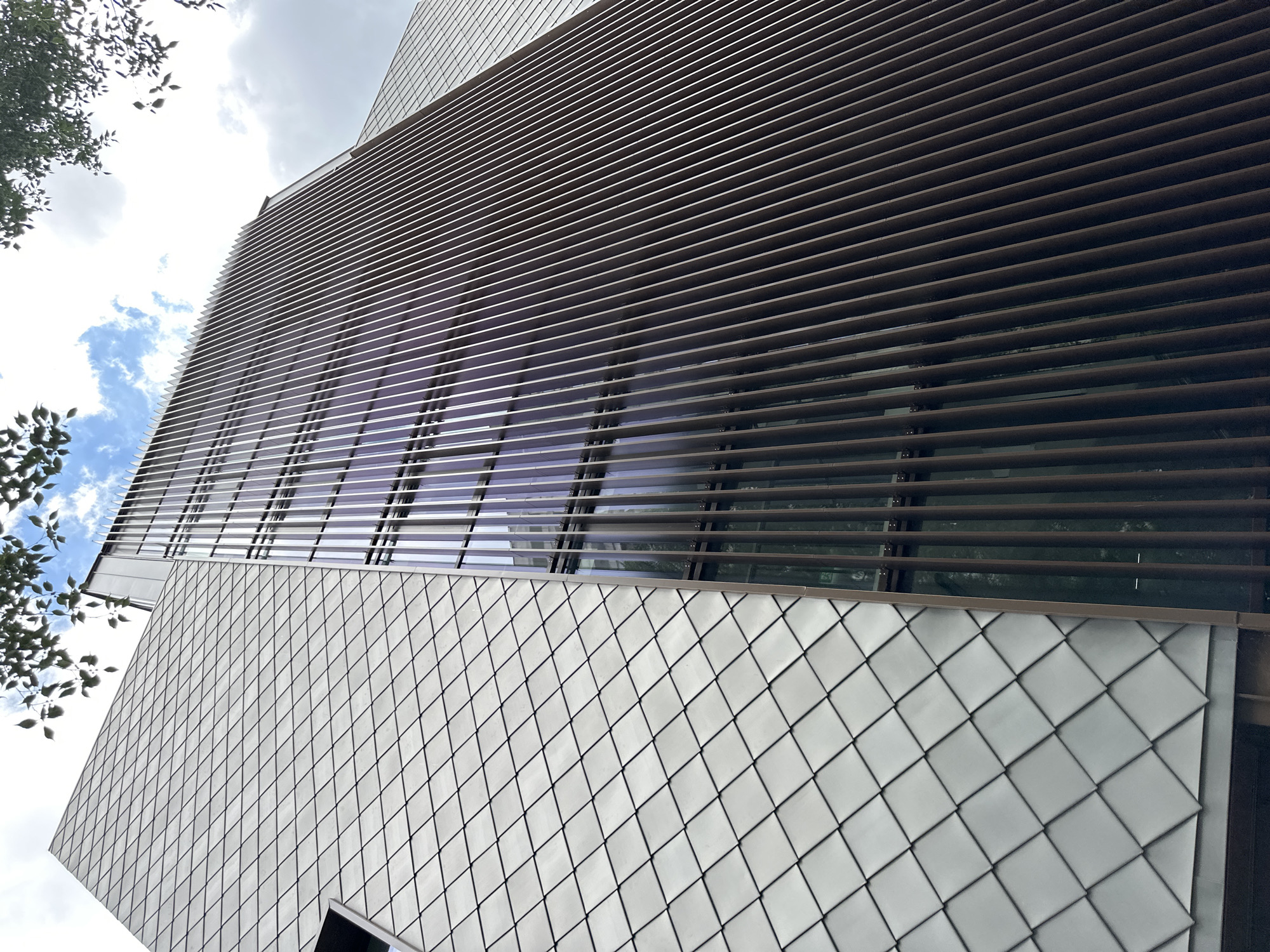 The image shows a low-angle, worm's-eye view of the exterior of a modern building with two distinct facade treatments. On the left, the wall is covered in diamond-shaped, metallic shingles. On the right, a large section of the building is clad in vertical, dark brown slats that appear to be metal or wood, creating a ribbed effect. Between these two sections, a portion of the building features floor-to-ceiling windows with a purplish tint, and the vertical slats are visible in front of these windows. The sky above is partly cloudy, with some blue patches visible. Tree branches with green leaves overhang the top of the building on the left side. The building's architecture suggests a contemporary urban setting. This building is located in Sofia, Bulgaria.