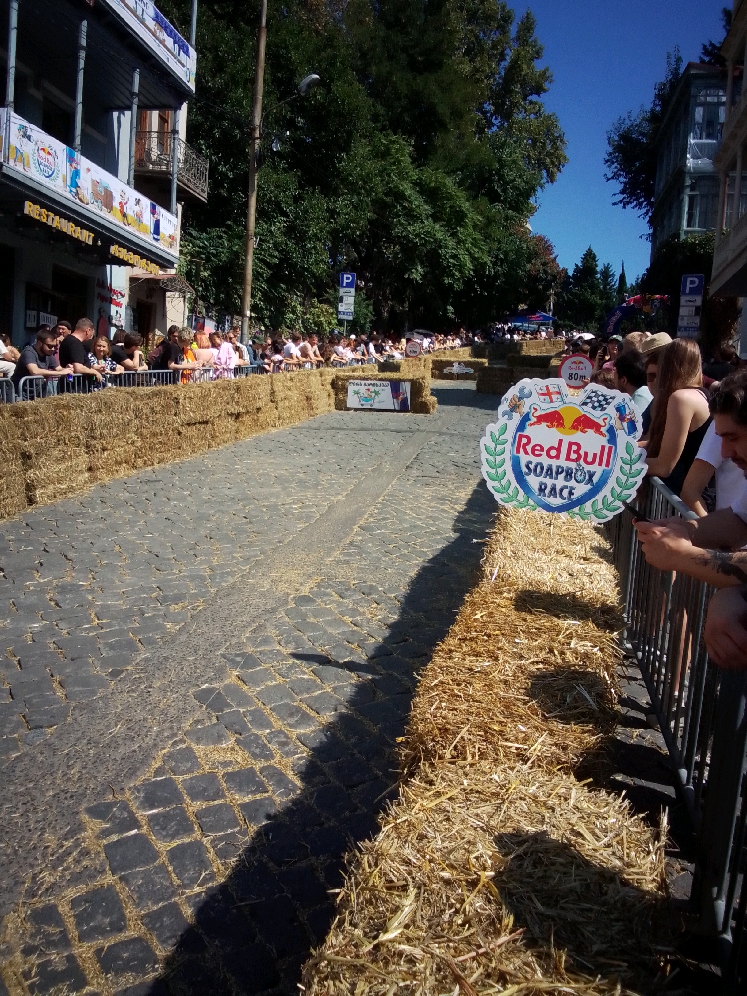 The image depicts a Red Bull Soapbox Race event taking place on a cobblestone street in Tbilisi, Georgia. A large crowd of spectators lines the street, separated from the racecourse by metal barriers and bales of hay. The buildings lining the street appear to be old, with balconies and ornate architectural details, suggesting a historical area of the city. One building in the background has a banner advertising "Restaurant," and another has a "P" sign indicating parking. The sky is clear and blue, with lush green trees visible behind the buildings. Several Red Bull logos and race-related signage are present, including a prominent Red Bull Soapbox Race sign on a hay bale. People in the foreground are watching the race, some holding phones to capture the event. The overall atmosphere suggests a lively outdoor event in a vibrant urban setting.